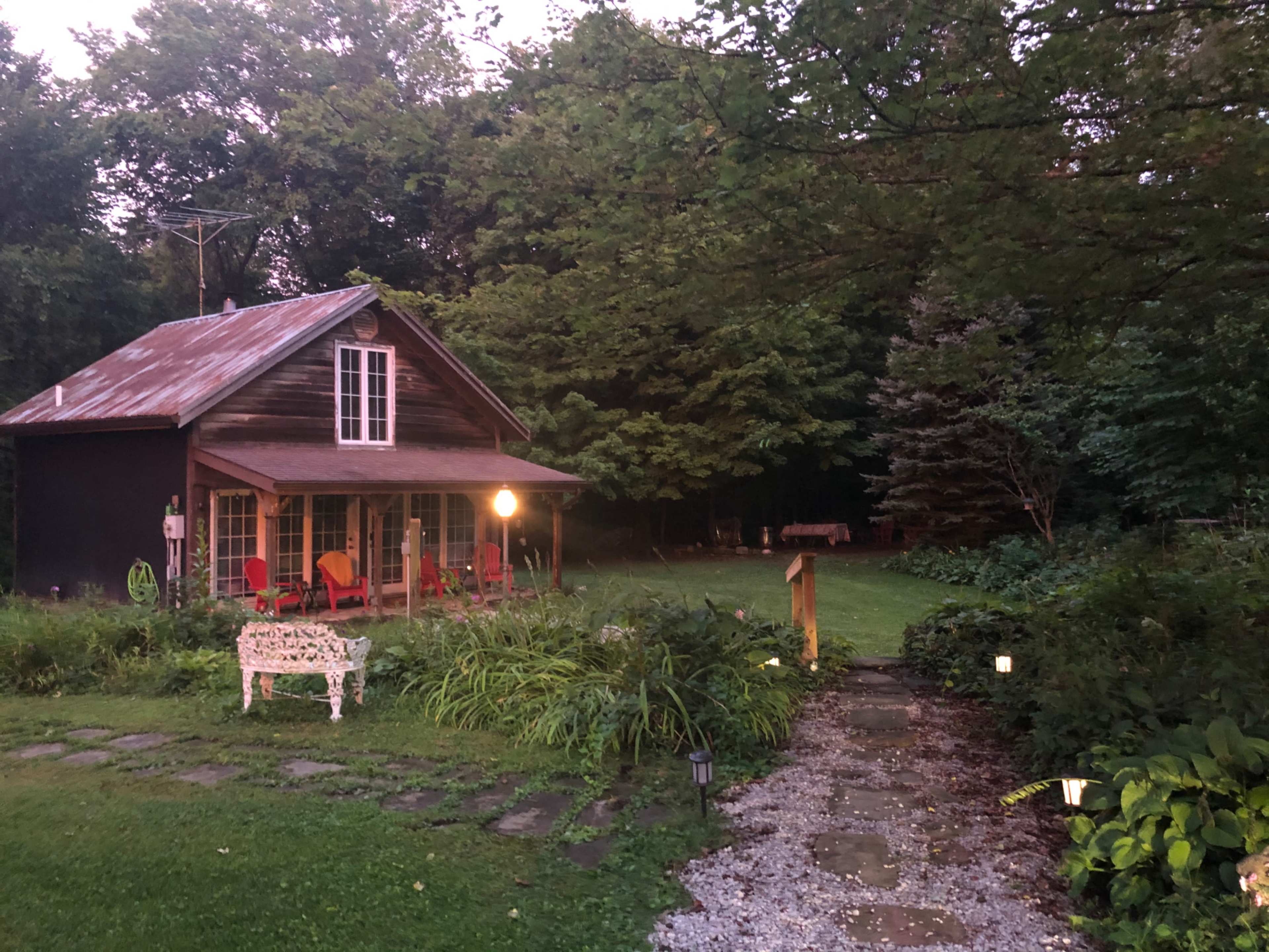 A two-story wooden house with a metal roof is set in a garden surrounded by trees, with a pathway leading to the front porch furnished with red chairs.