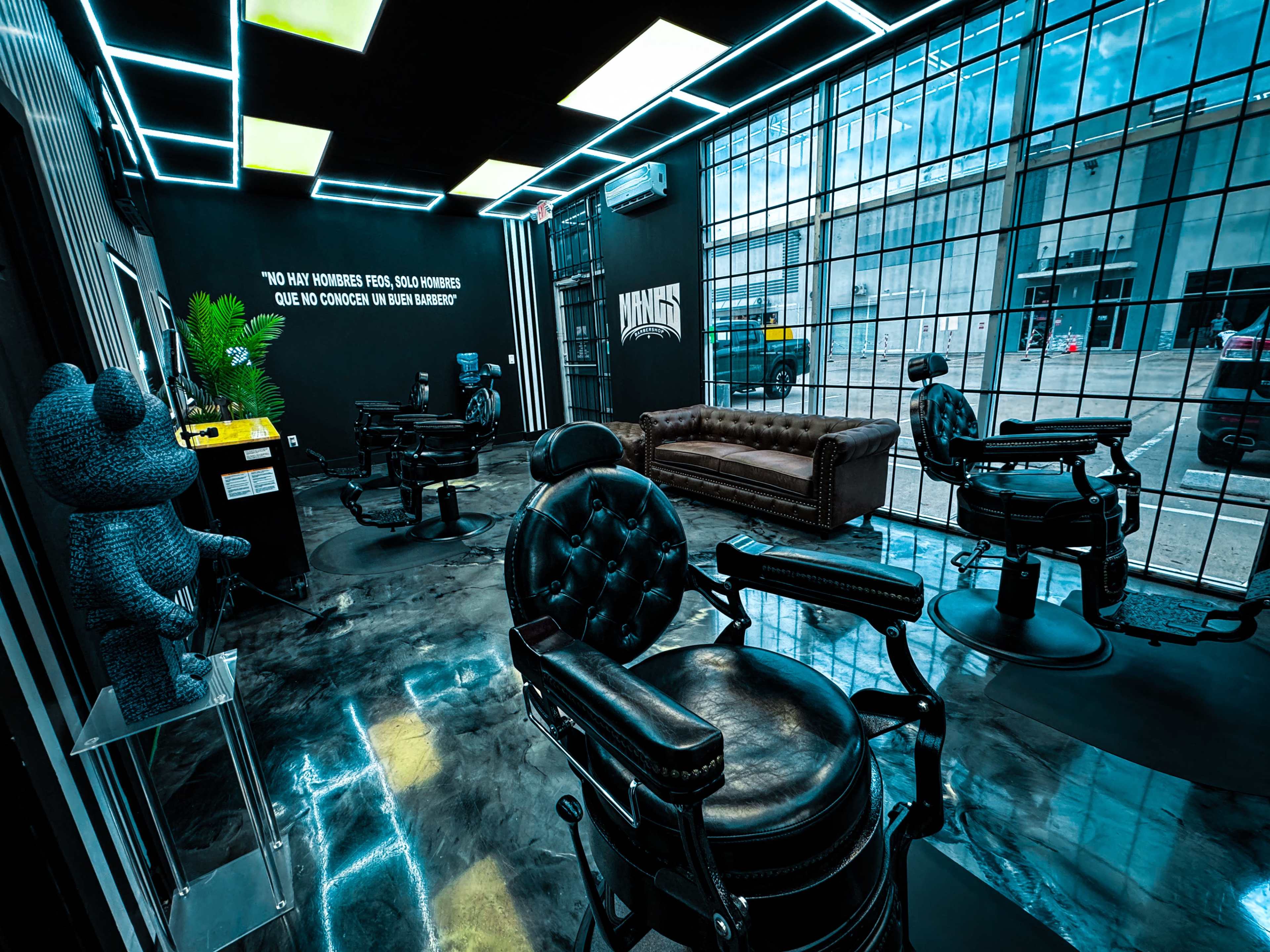 The interior of a modern barbershop features black leather barber chairs, a plush brown sofa, and large windows with grid patterns.