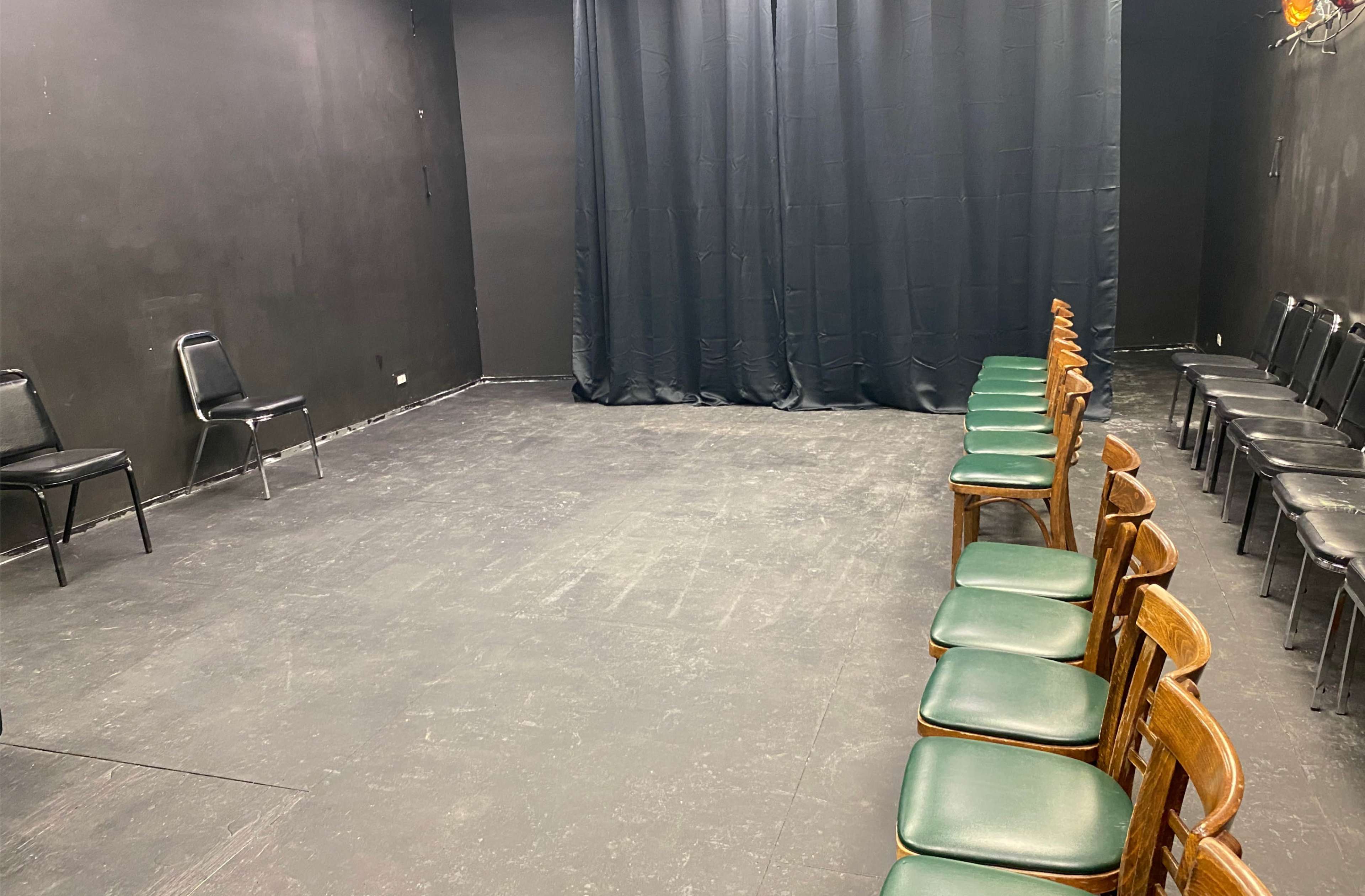 The image shows a small black-walled theater space with two rows of chairs facing a curtain.
