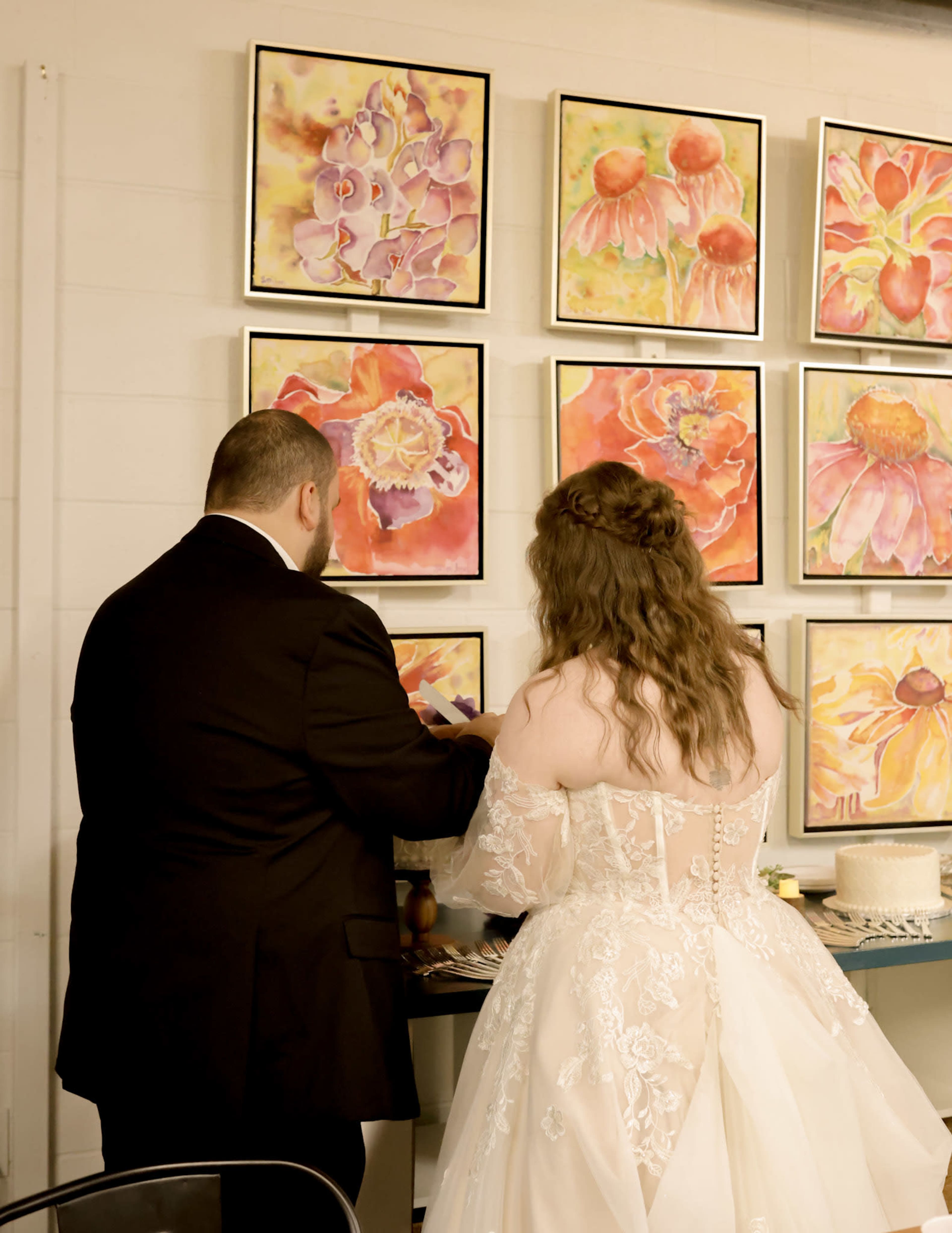 A couple stands together, with the man in a black suit and the woman in a lace wedding dress, facing a wall adorned with colorful floral paintings.