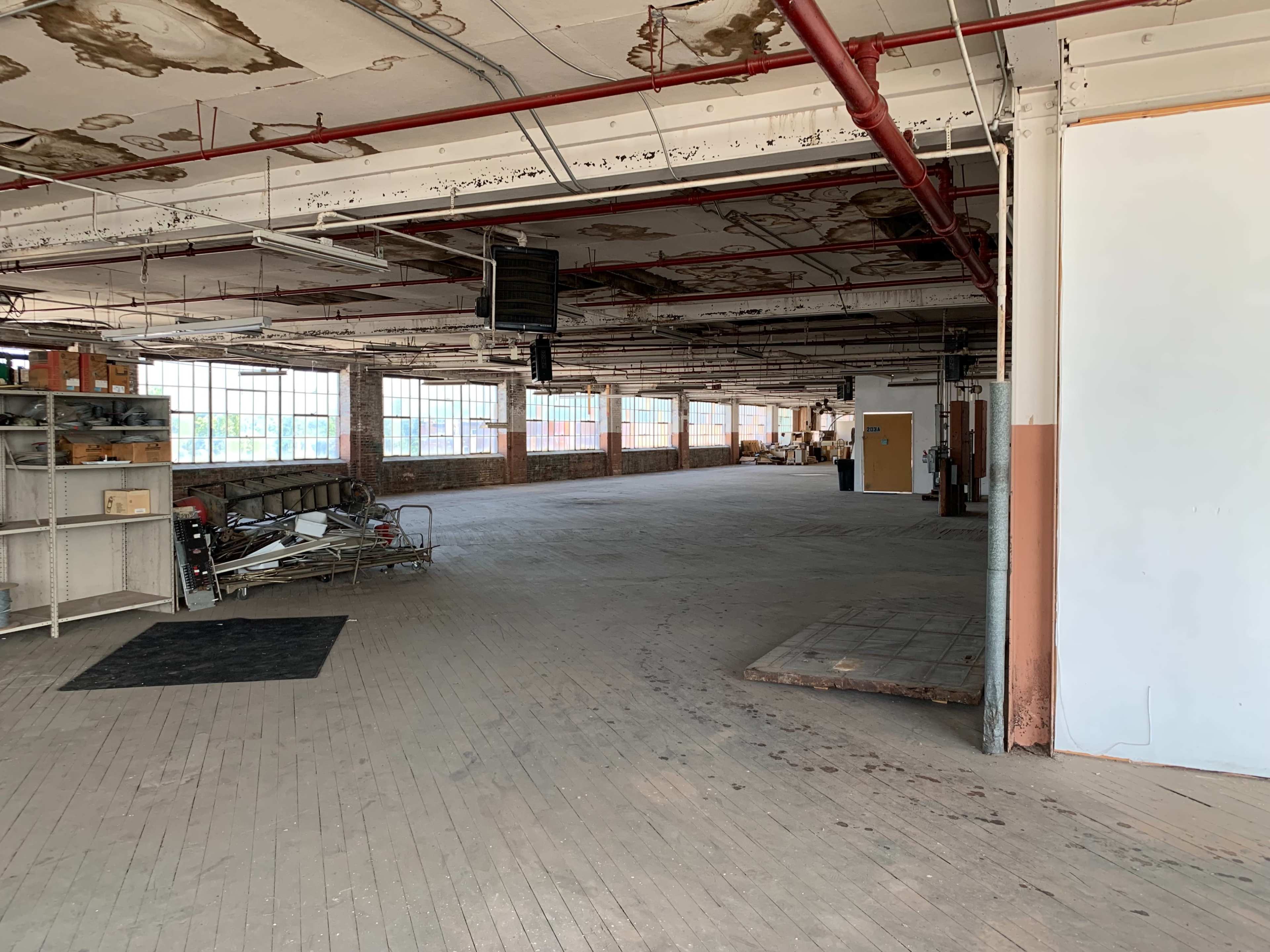 The image shows a spacious, vacant industrial interior with large windows and exposed ceilings.