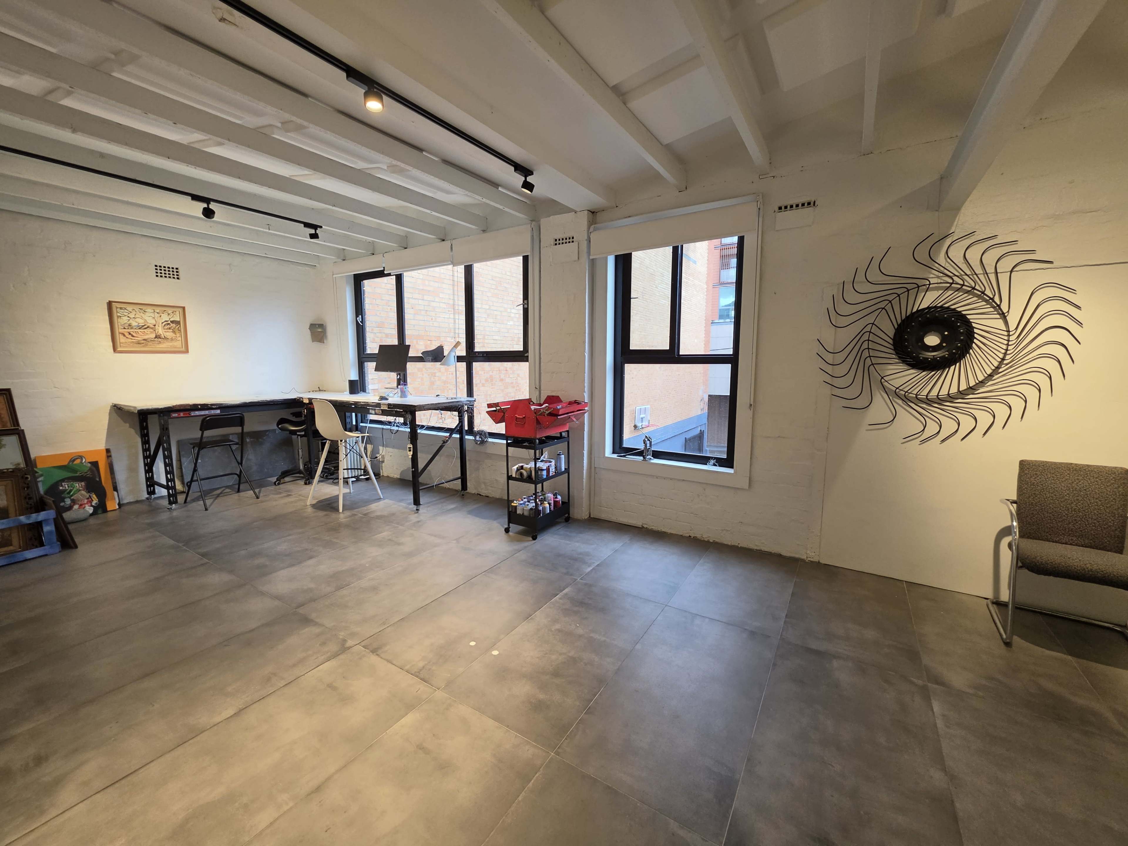 The image shows a spacious, well-lit room with large windows, a desk and chair setup, shelves with red bins, and a decorative wall feature.