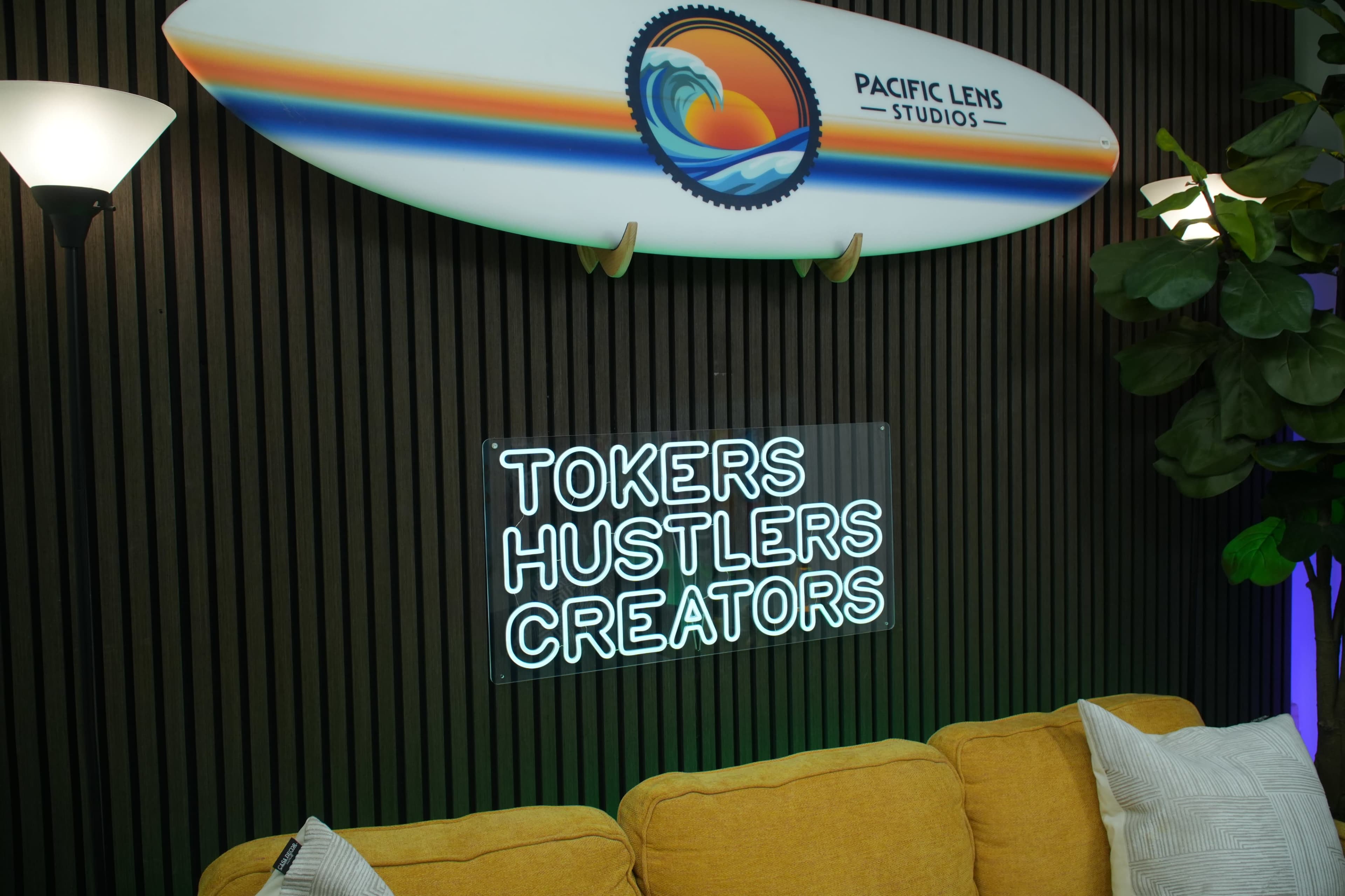 A yellow couch is positioned under a neon sign that reads "TOKERS HUSTLERS CREATORS," with a surfboard hanging above it.