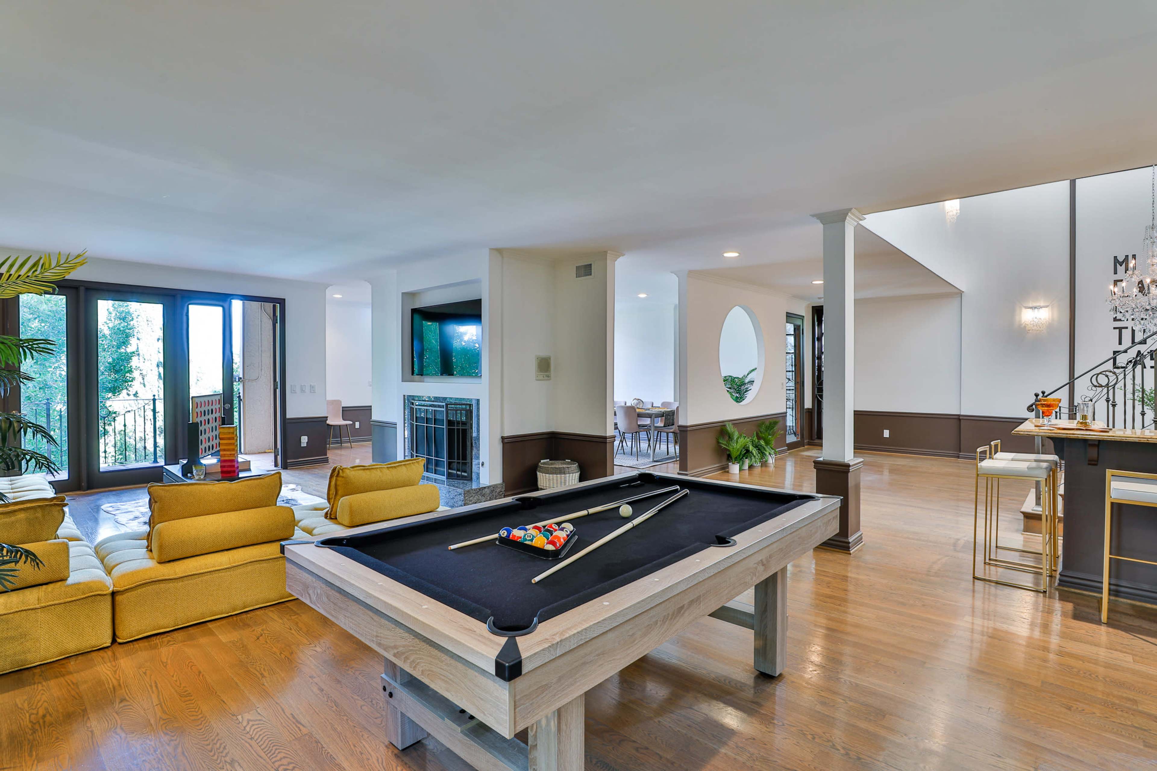 A spacious room featuring a pool table at the center, with a seating area and a dining space visible in the background.