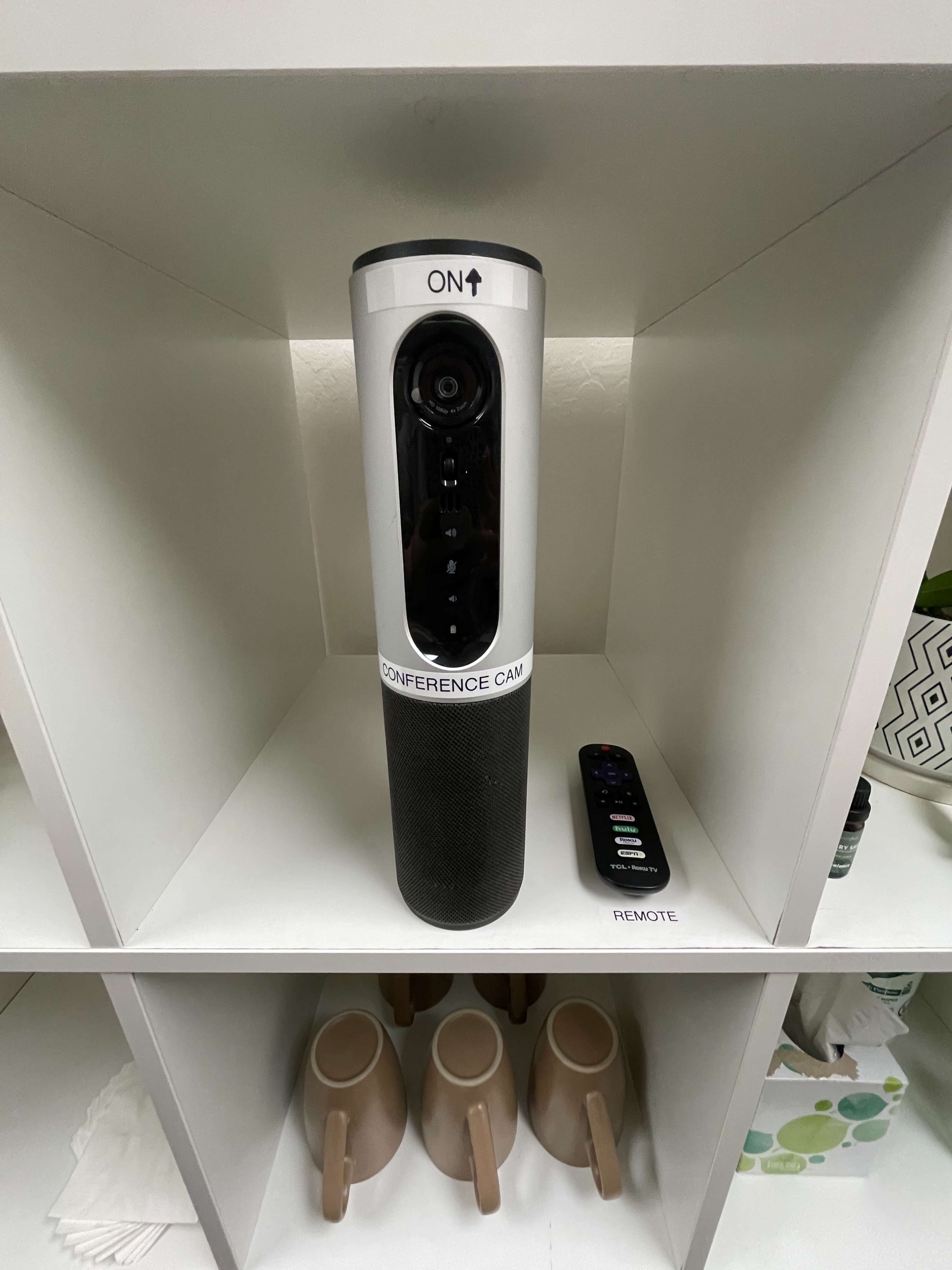 A conference camera with a remote is displayed on a shelf among ceramic mugs and decorative items.
