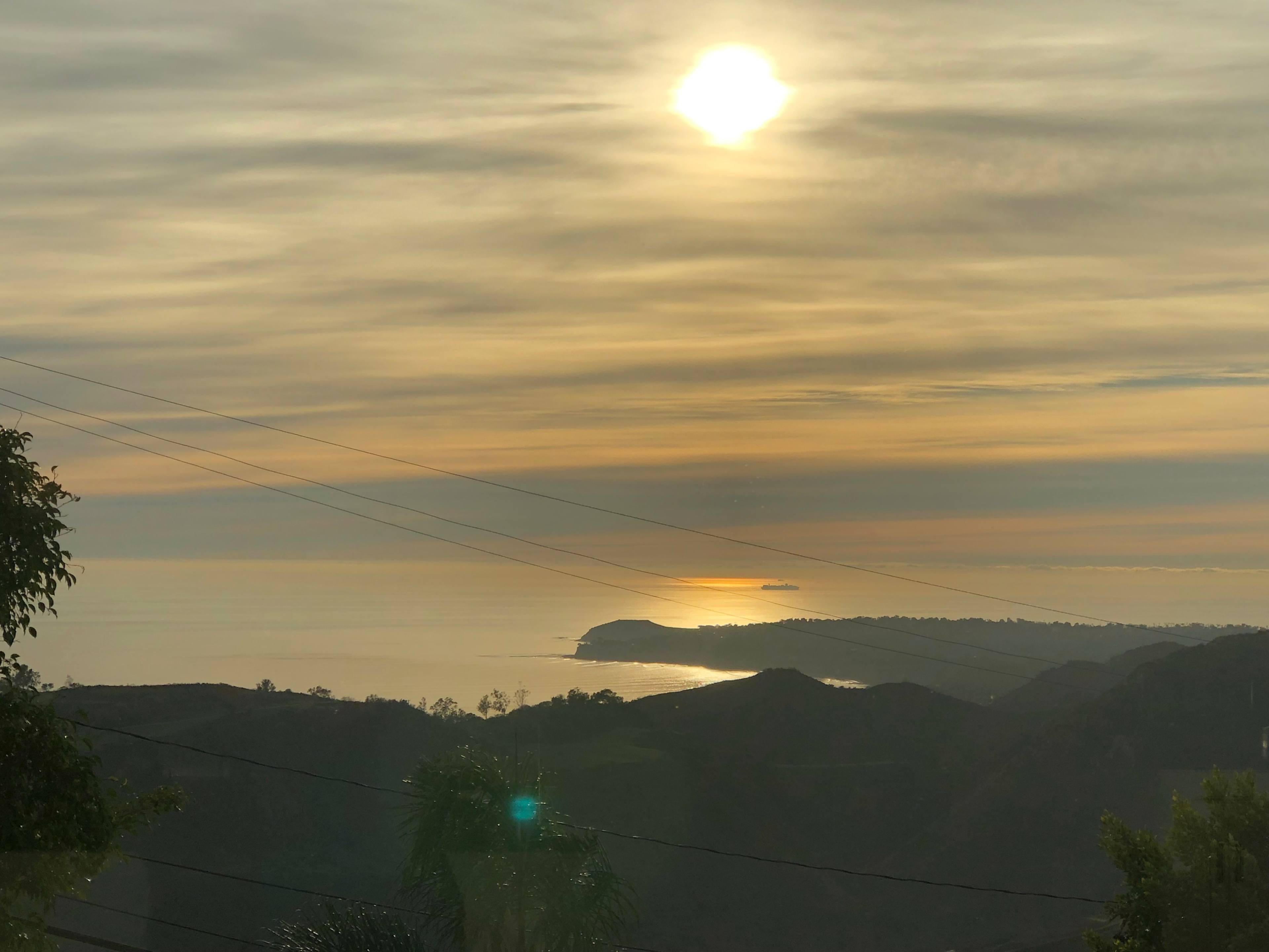 A sunset over a calm ocean, with a faint outline of a distant shoreline and silhouetted hills in the foreground.