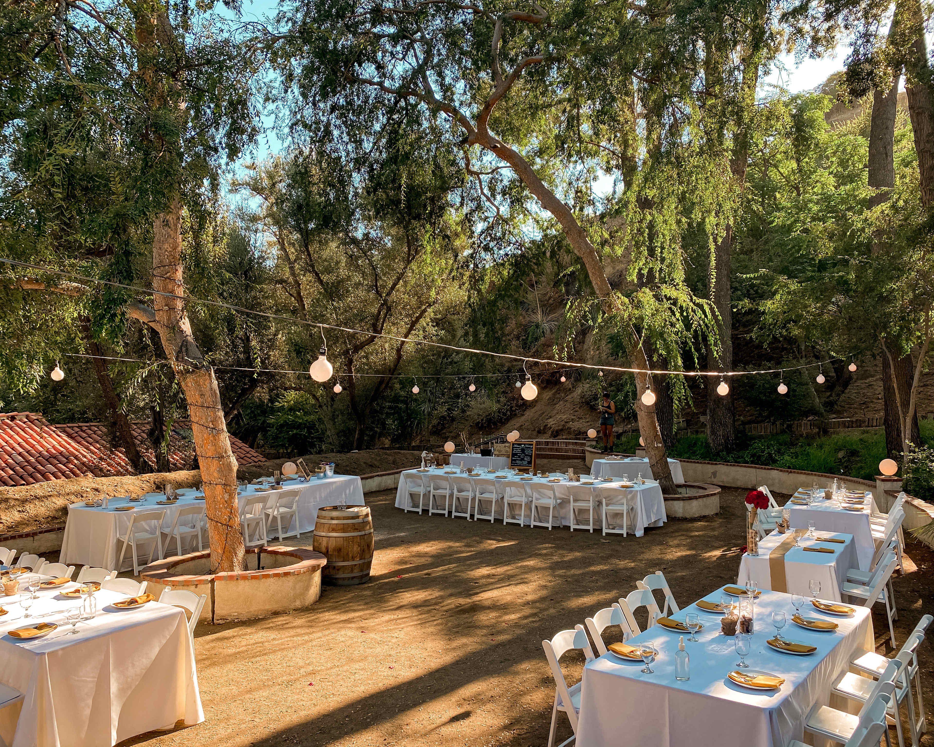 A dining area with white tables and chairs is set up outdoors under trees, adorned with string lights.