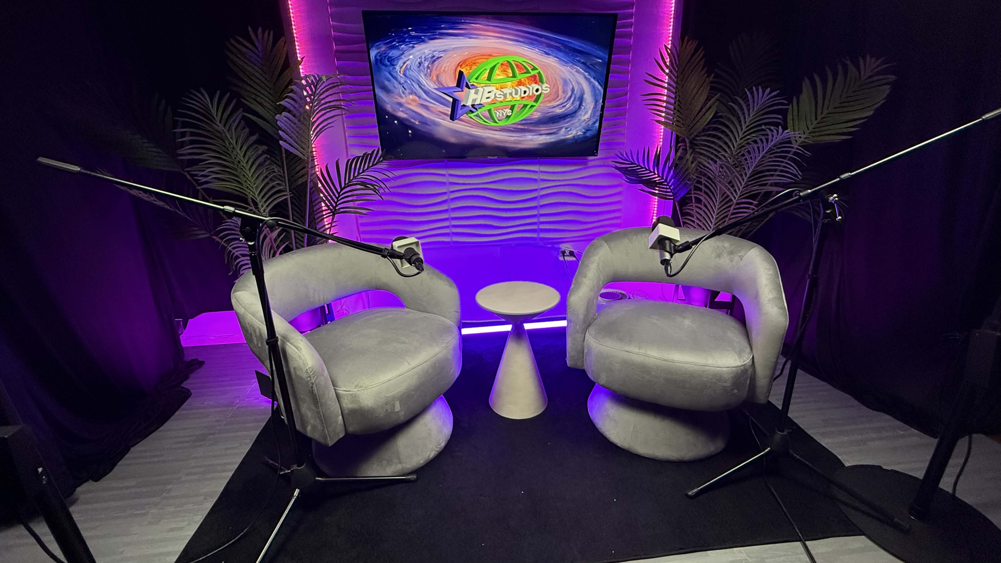 The image shows a podcast recording studio featuring two gray chairs, a small white table between them, and a screen with colorful graphics in the background, illuminated by pink lights.