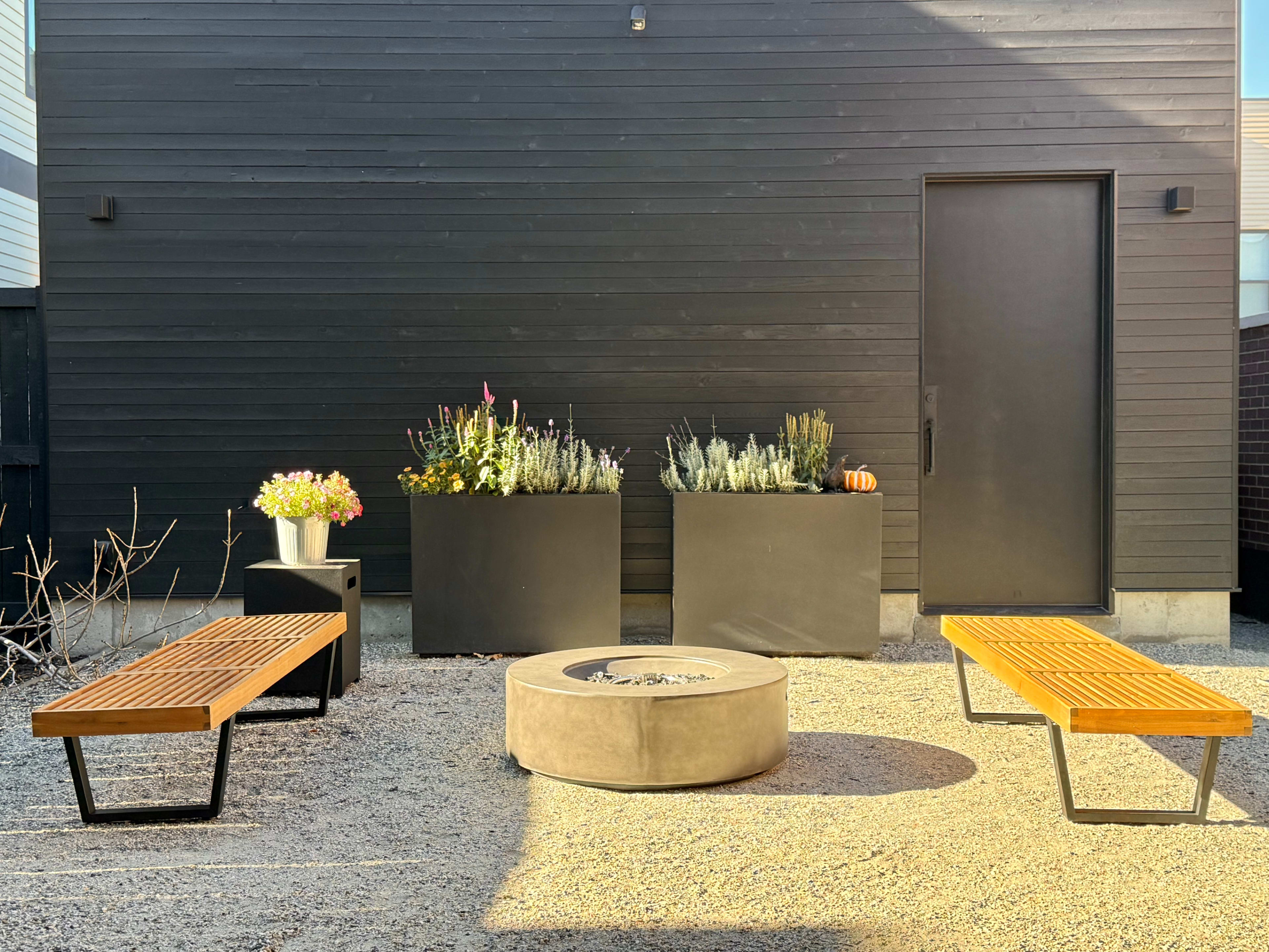 The image features a patio with two wooden benches, a circular fire pit, and planters filled with various plants positioned against a black wooden wall.