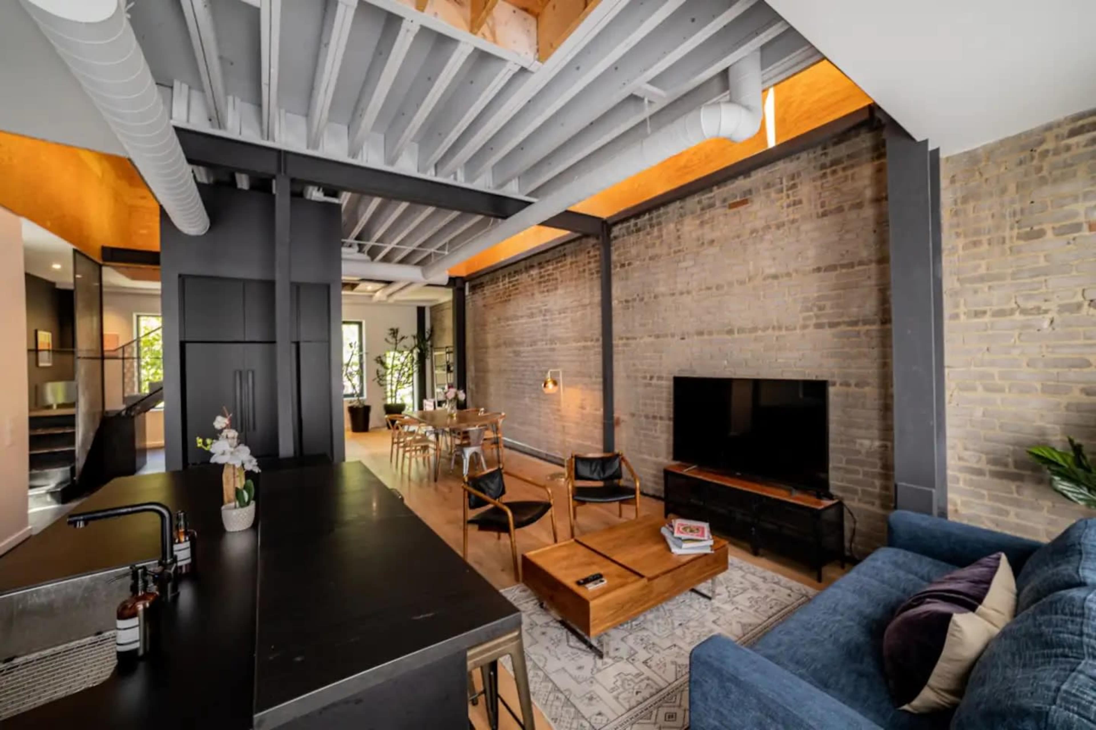 An open living space with a modern kitchen, dining area, and a comfortable seating arrangement, featuring exposed brick walls and wooden beams.