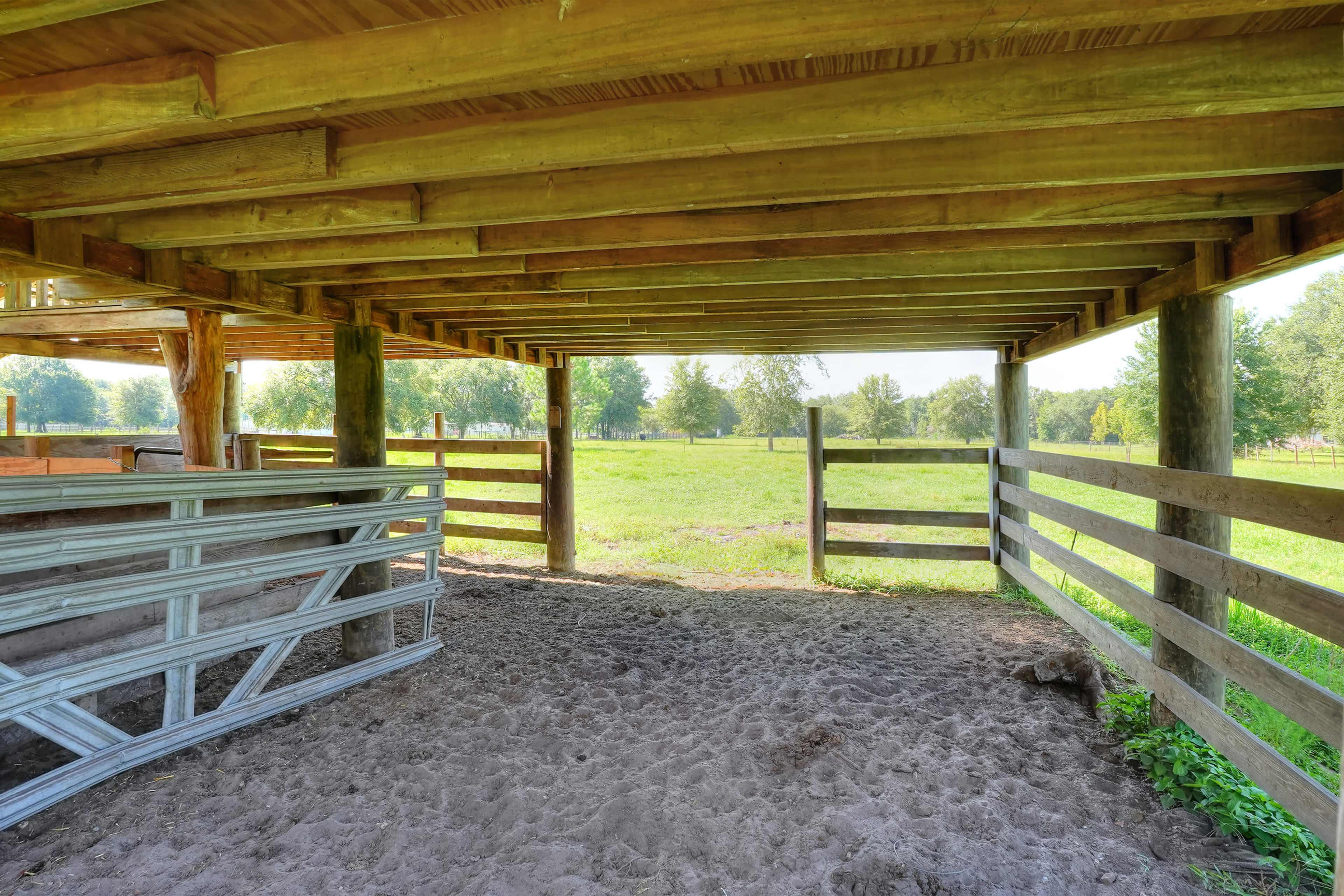 The image shows a shaded area beneath a wooden structure with a sandy floor and surrounding wooden fencing, overlooking a grassy landscape.