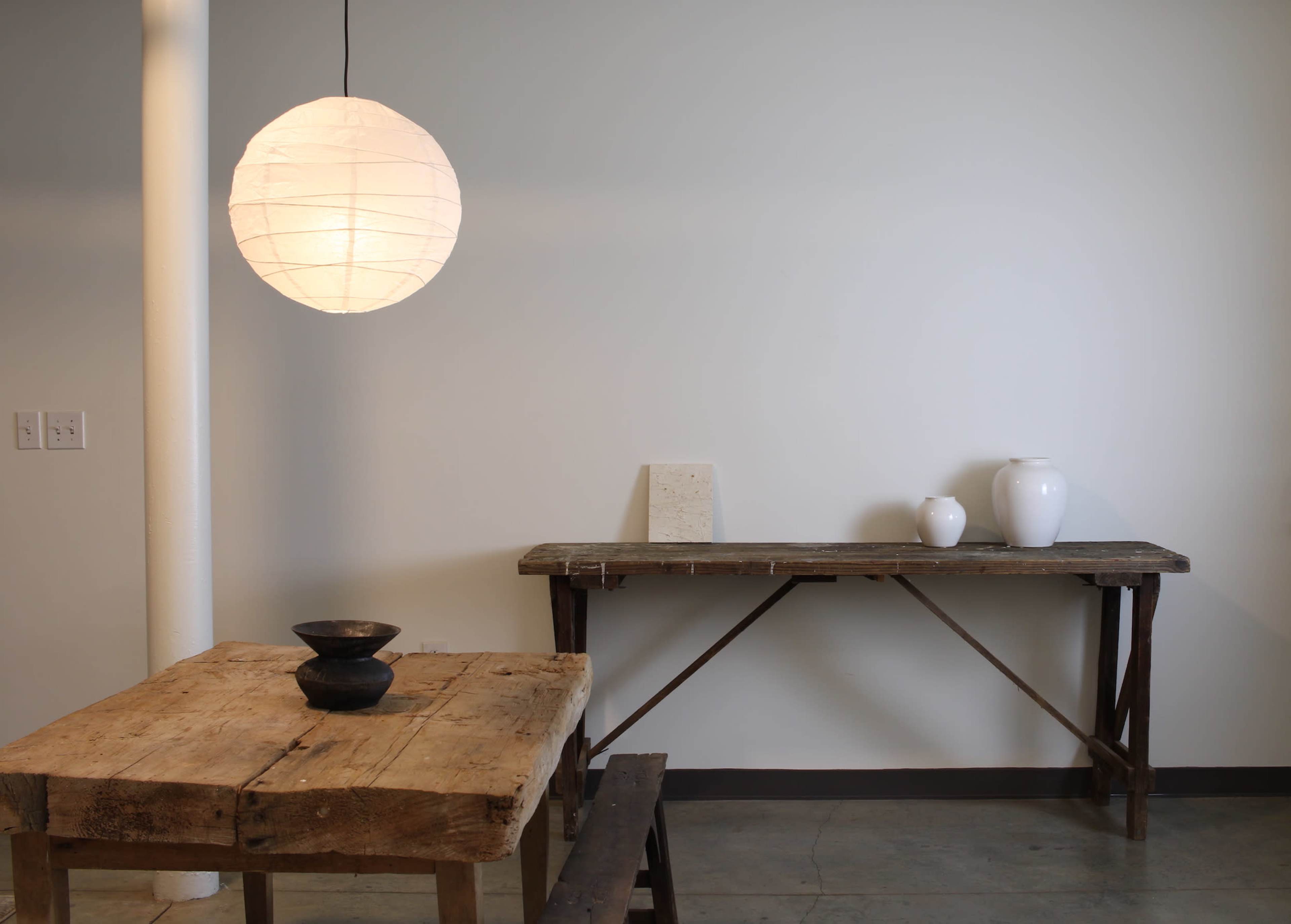 A wooden table and bench are positioned beneath a round pendant light, while a rustic shelf holds three white ceramic vases against a plain wall.