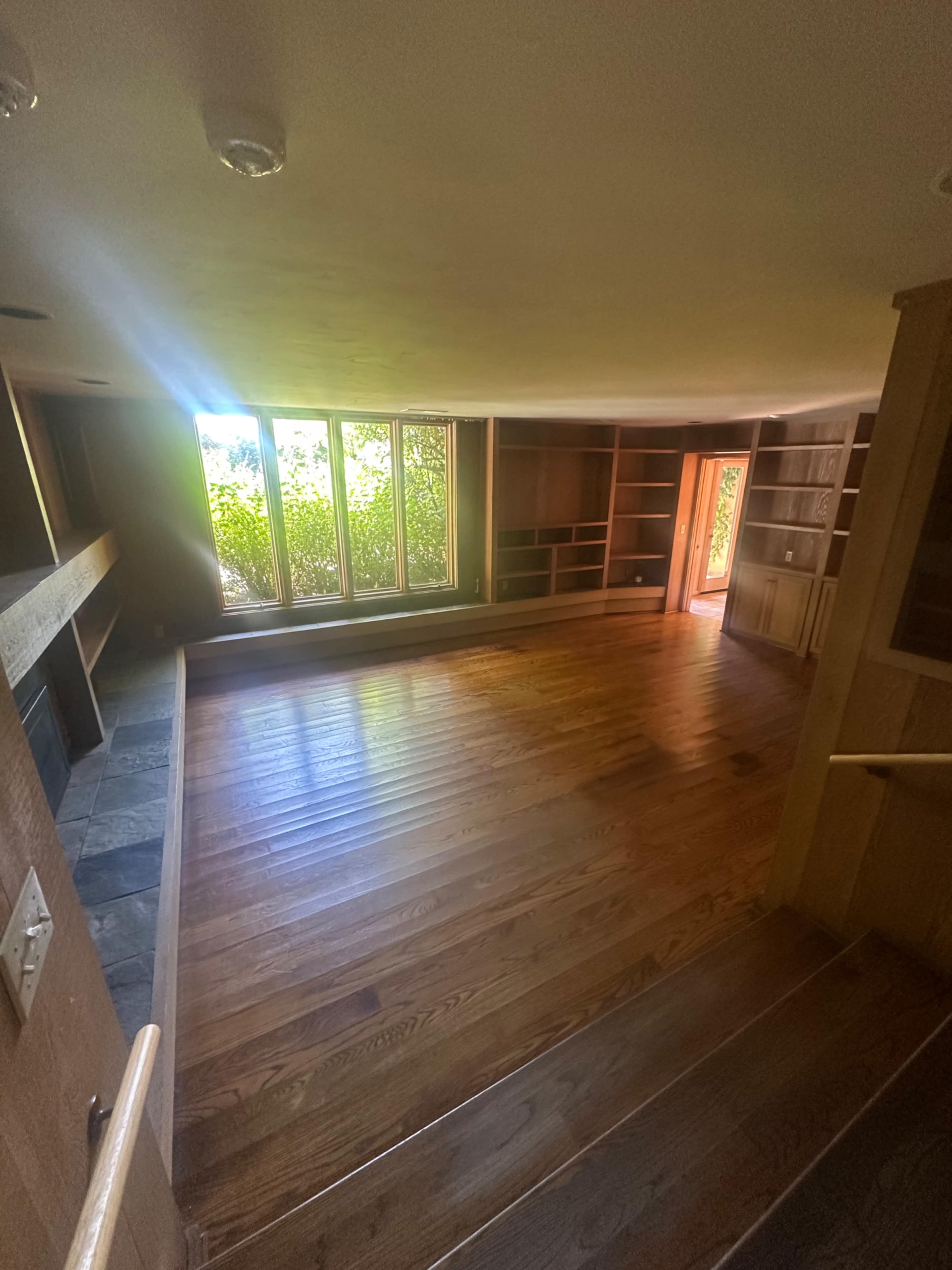 The image shows a spacious room with wooden flooring, large windows allowing natural light, and built-in shelves lining the walls.