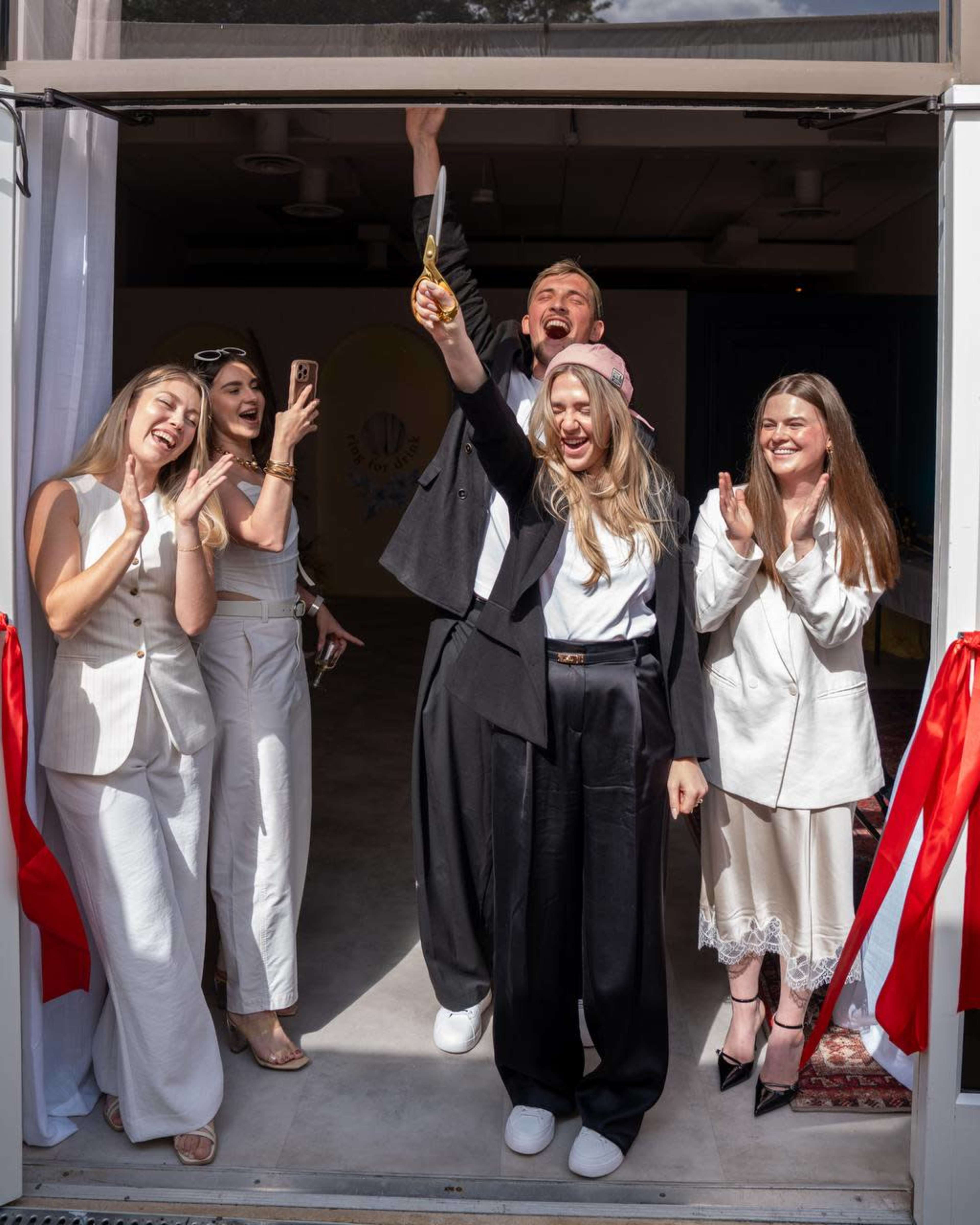 A group of five people stands in a doorway, celebrating as one person holds a pair of scissors with arms raised in triumph.