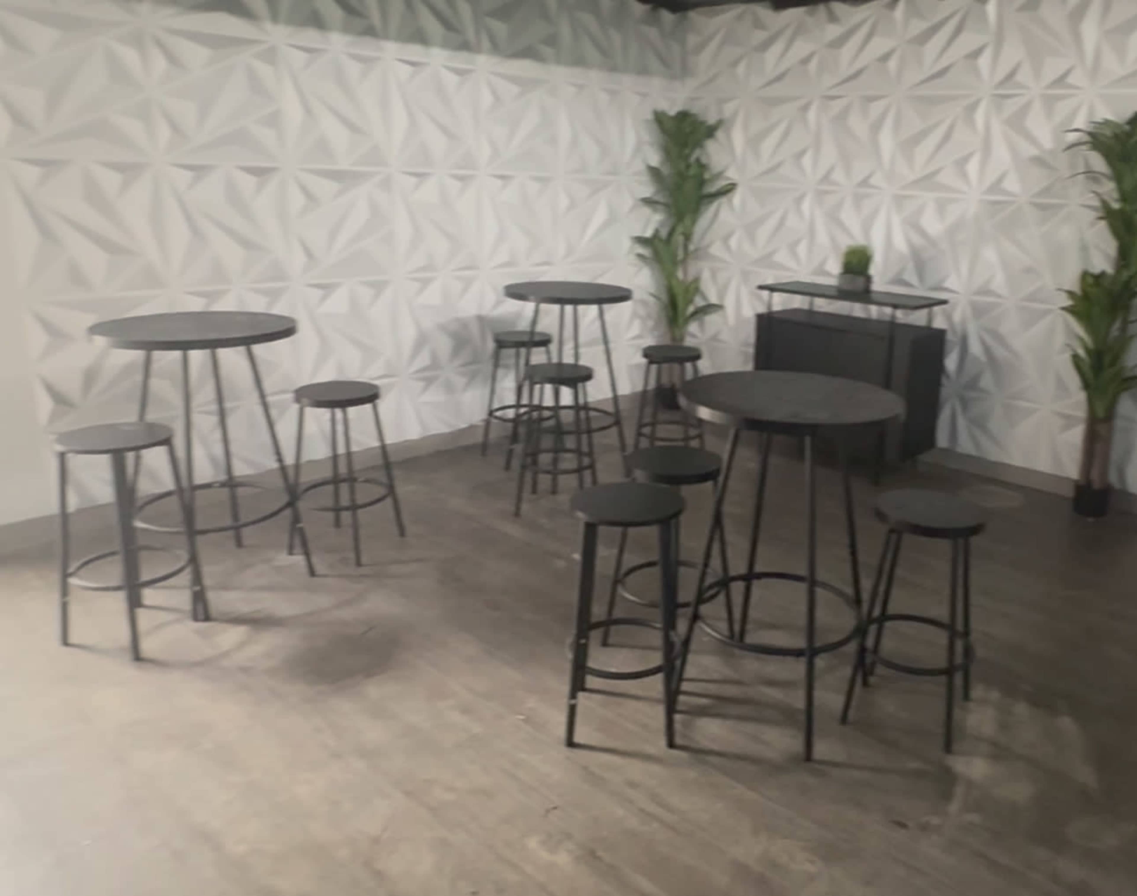 The image shows a modern venue with multiple round tables and stools arranged on a wooden floor, set against a textured white wall.