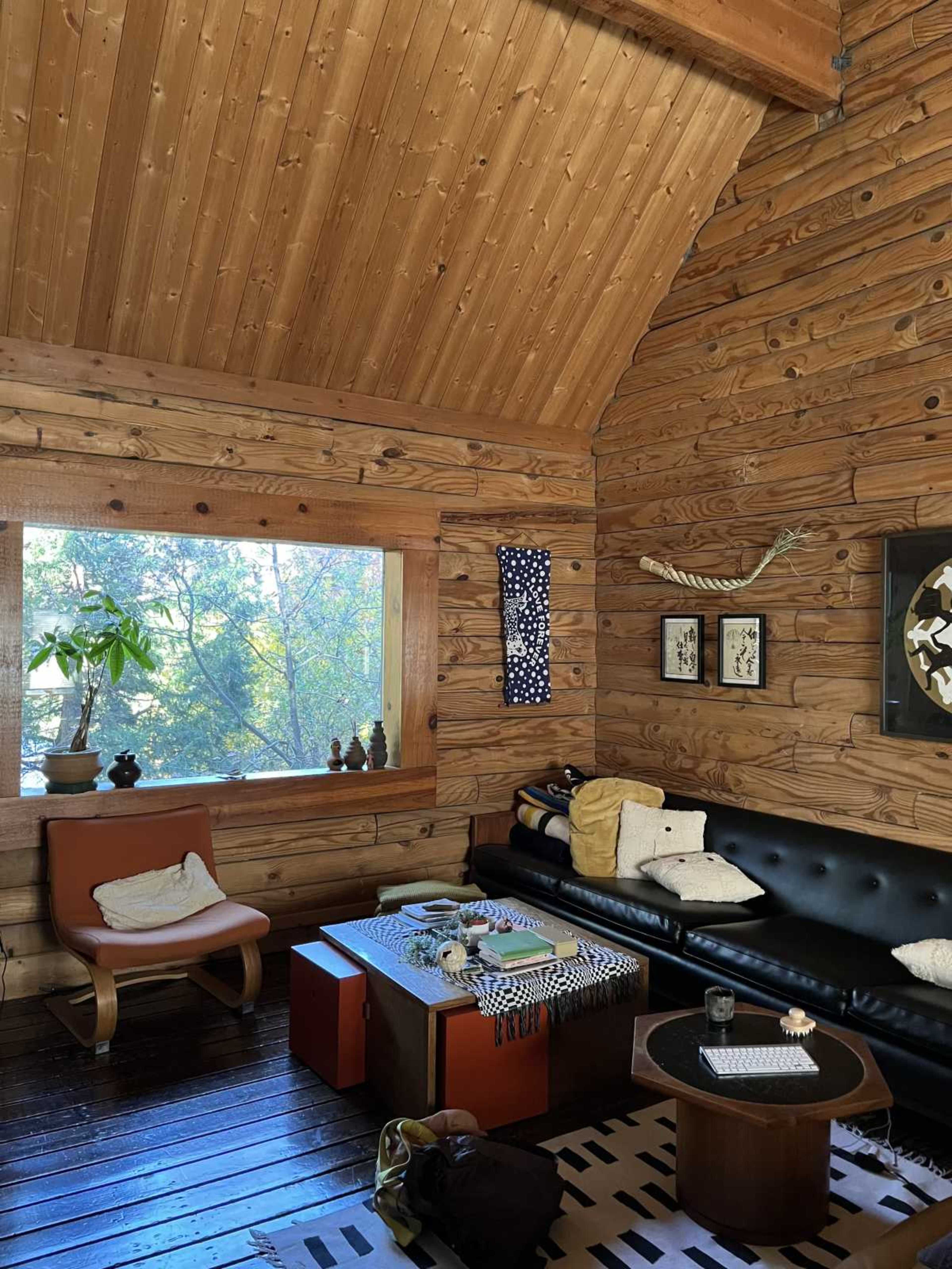 The image shows a cozy wooden living room with a large window, a black leather sofa, and various decorative items on the walls and surfaces.