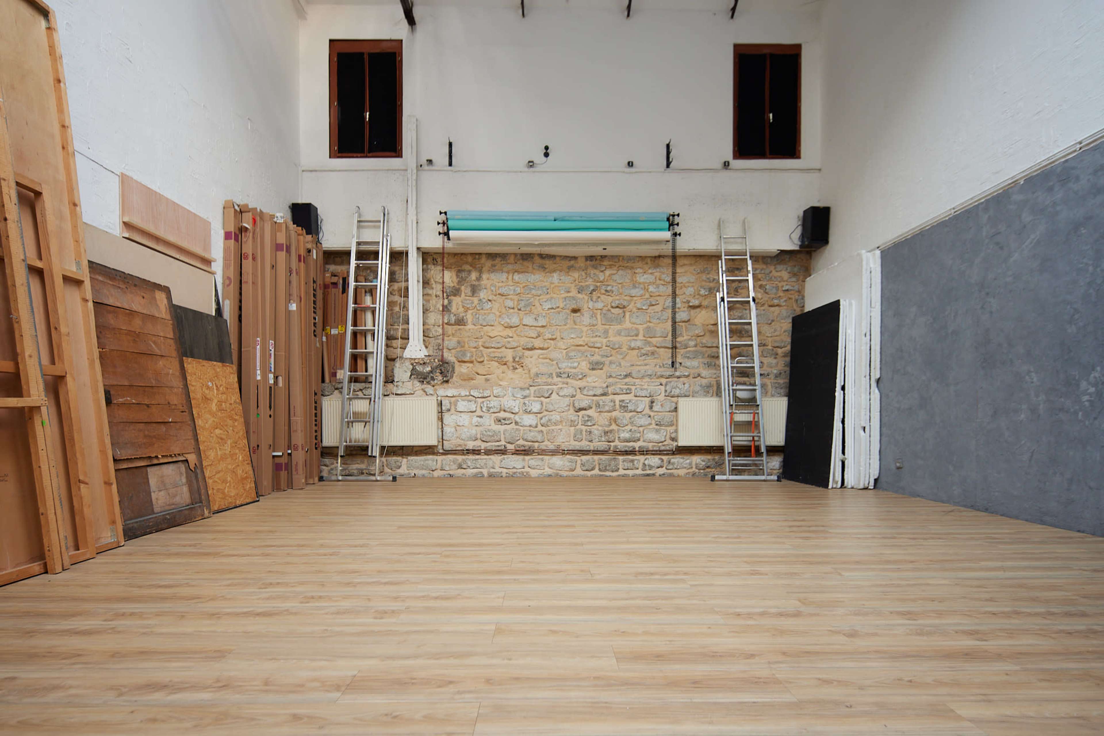 A spacious, empty room with wooden flooring, a stone wall at the back, and two ladders positioned on either side.