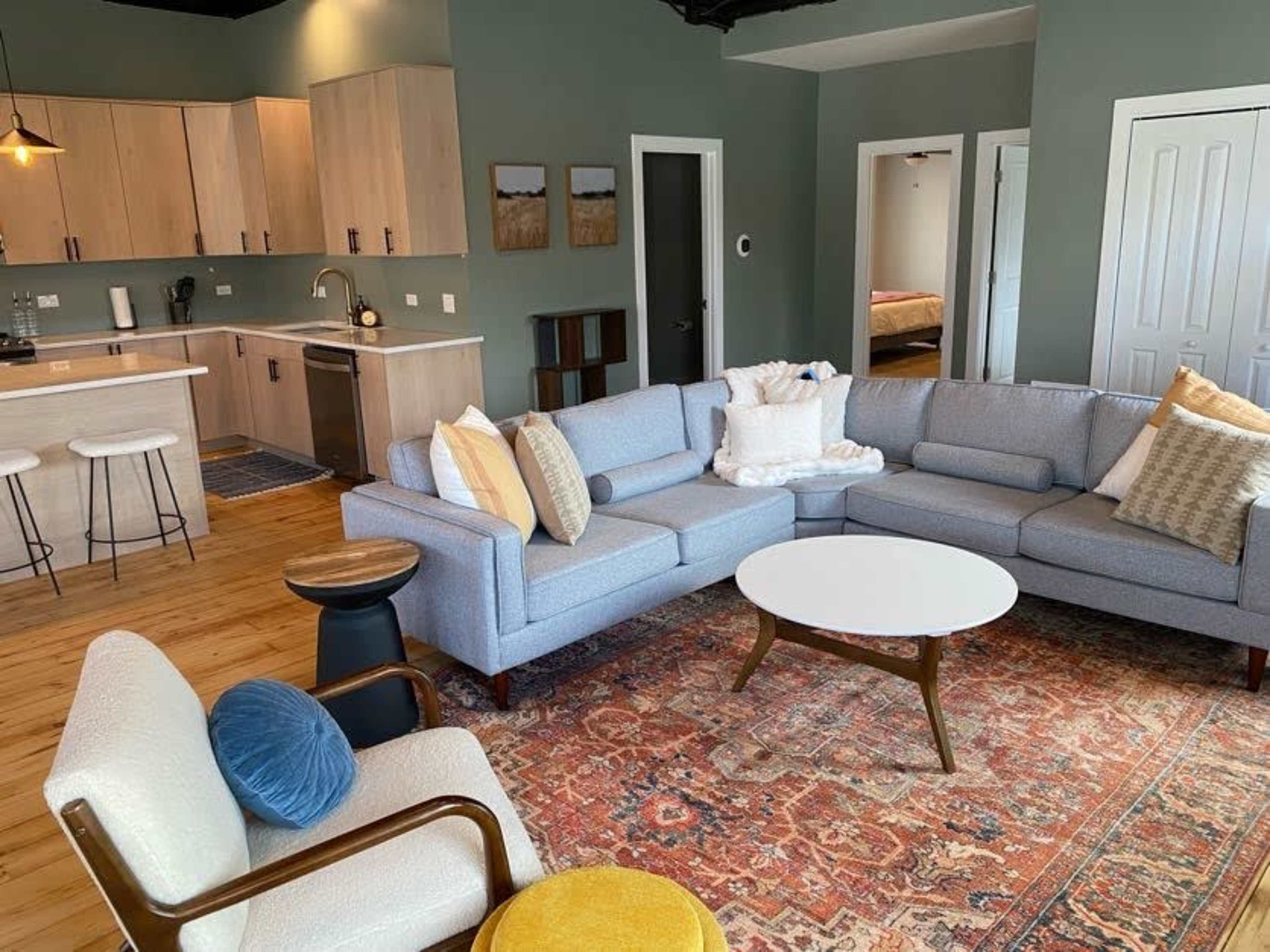 A modern living room featuring a light gray sectional sofa, a round coffee table, and wooden floors, with an open kitchen visible in the background.