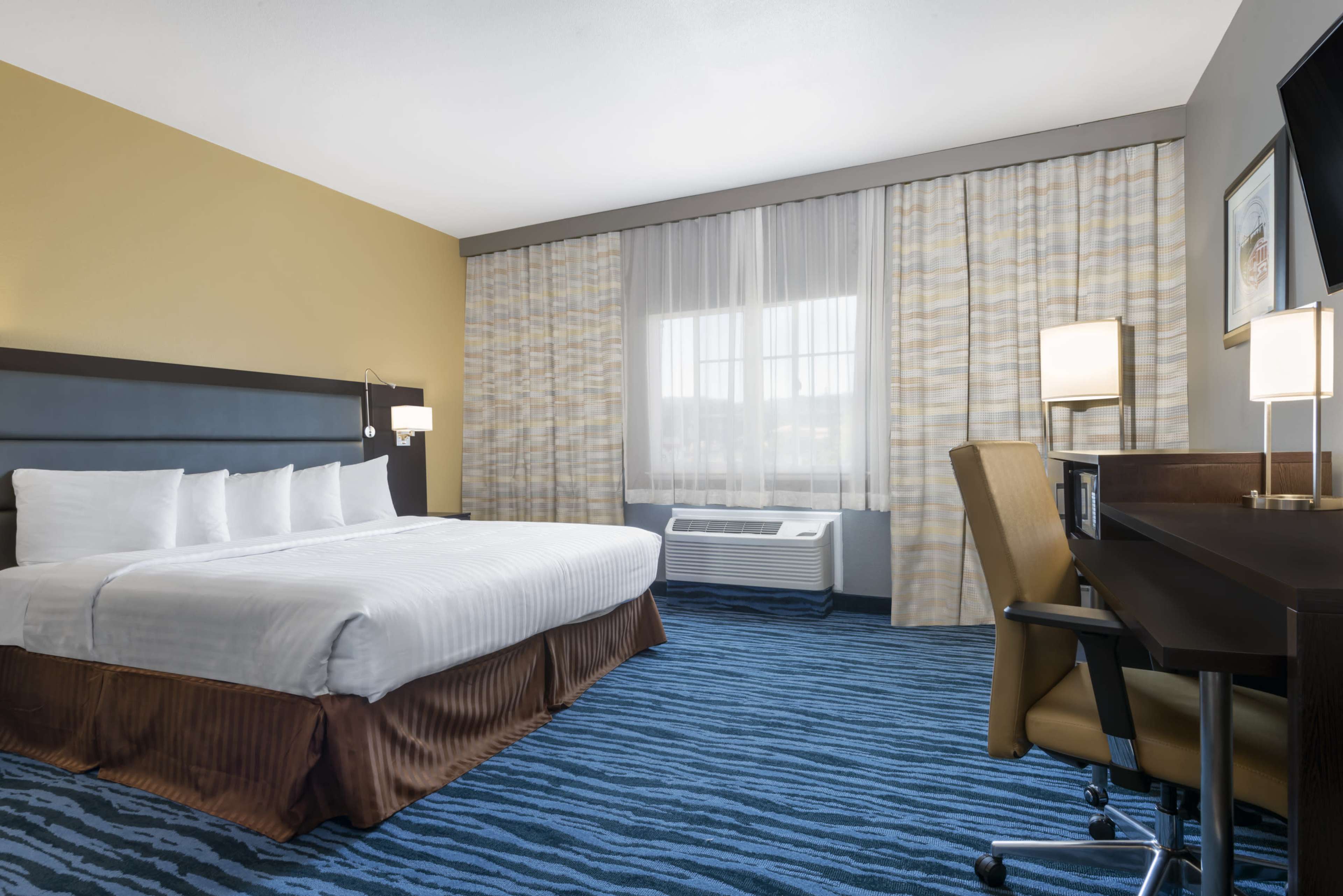 A hotel room with a queen-sized bed, a workspace with a desk and chair, large windows covered by curtains, and an air conditioning unit.