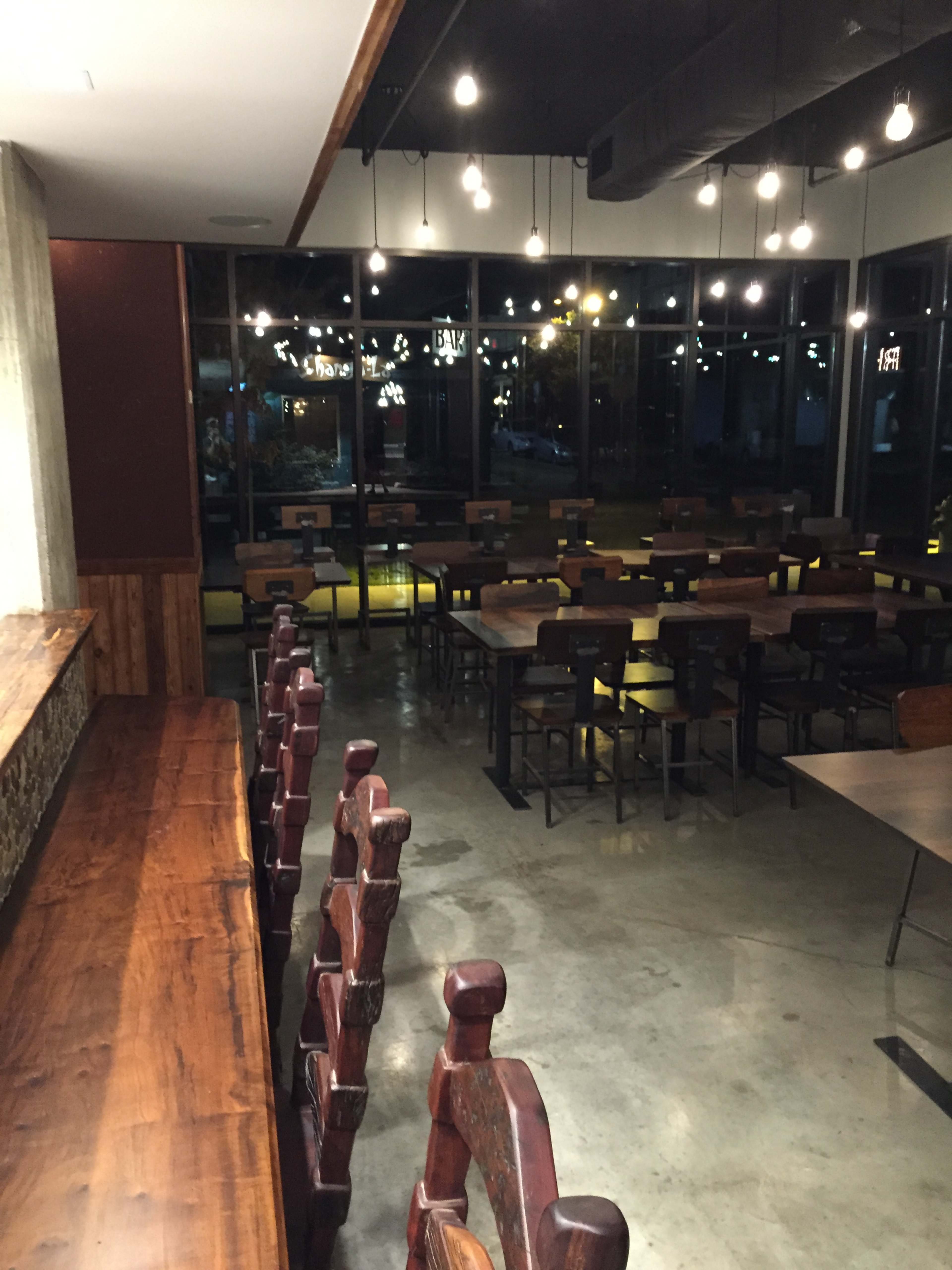 The image shows a dimly lit restaurant with wooden tables and chairs arranged around a polished concrete floor.