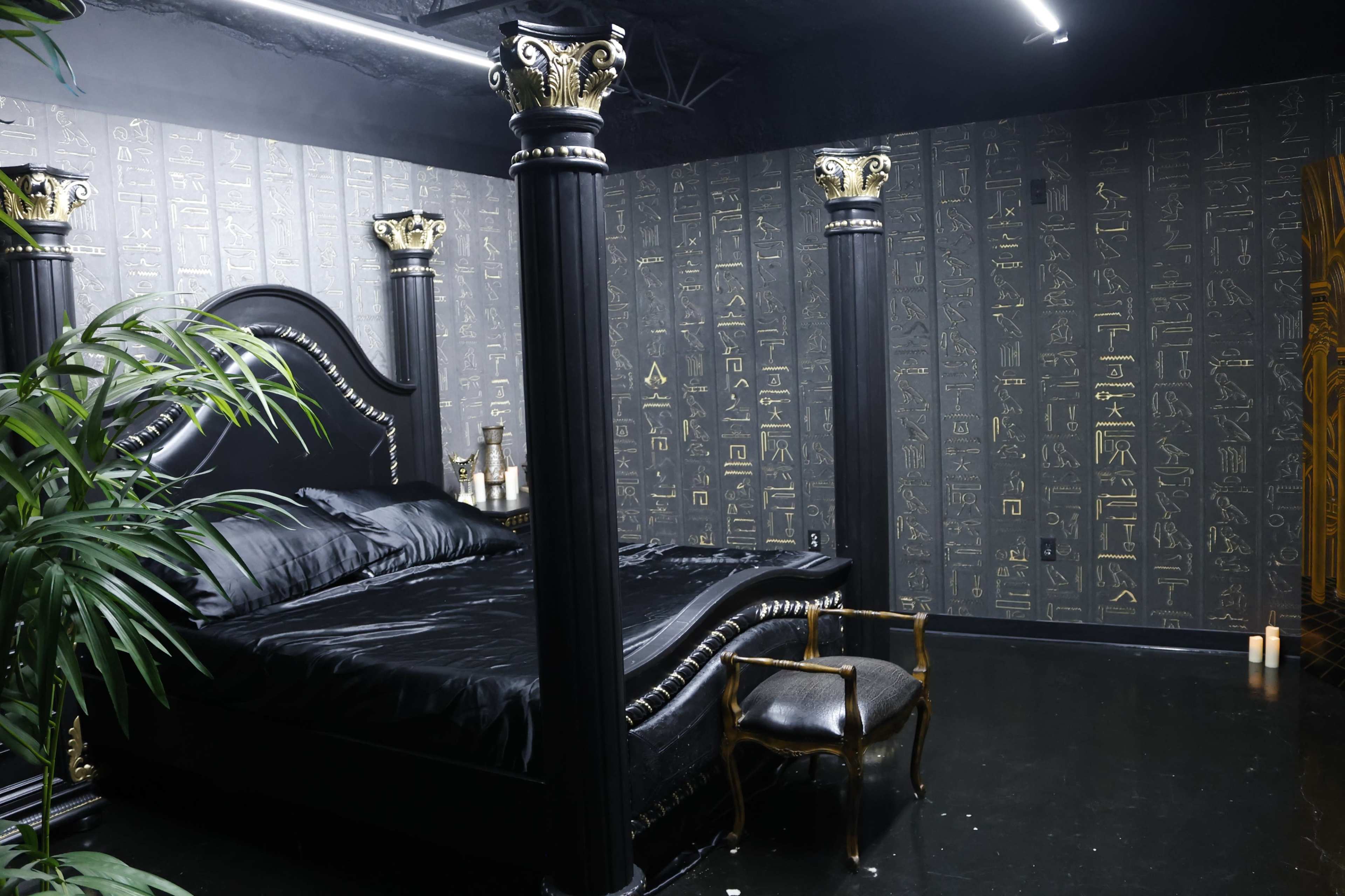 A dark room featuring a black bed with ornate columns and walls decorated with hieroglyphics.