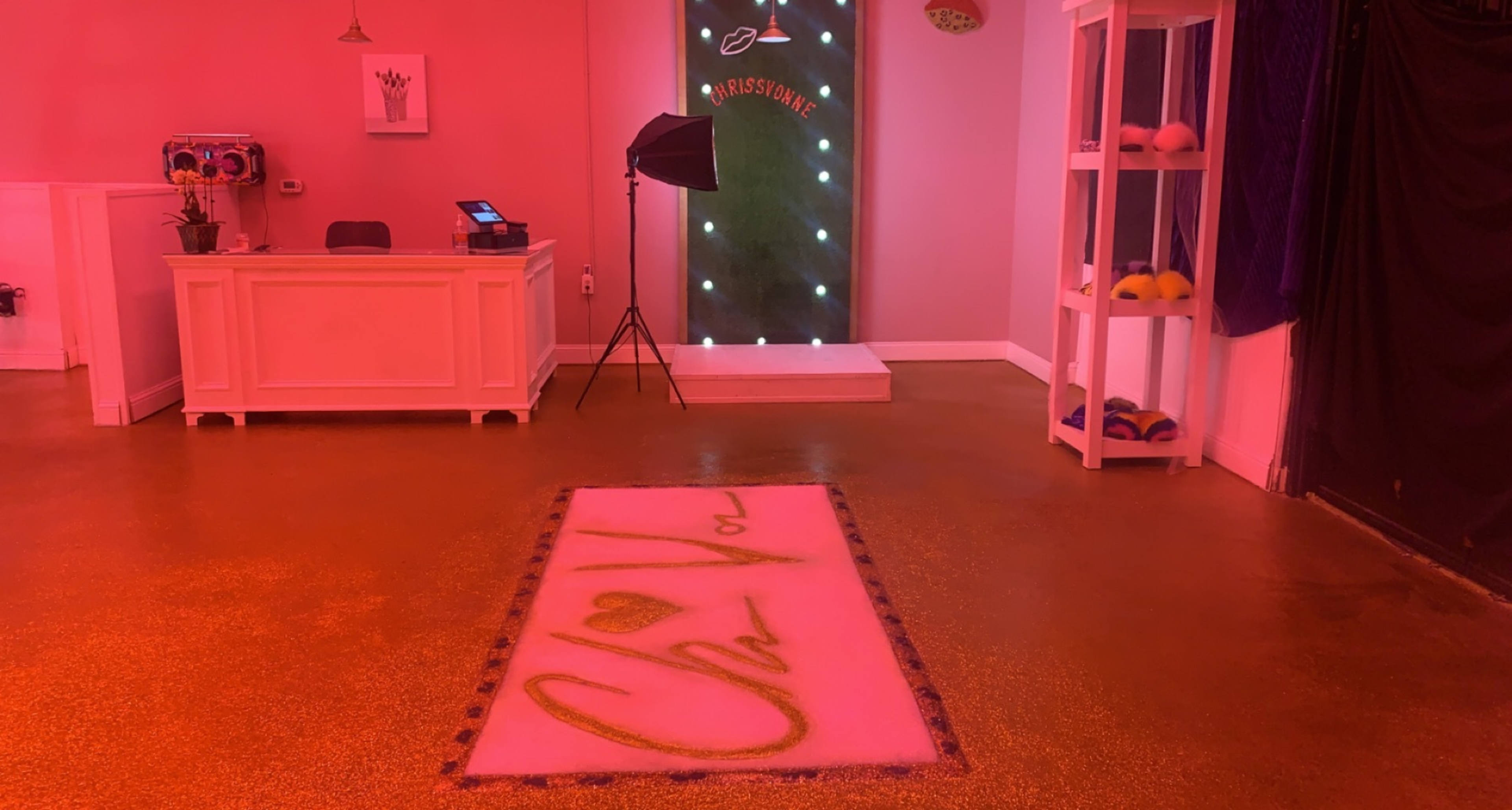 The interior of a brightly lit room features a welcoming reception area, a step-and-repeat backdrop, and a plush pink rug with the words "Oh You" prominently displayed.