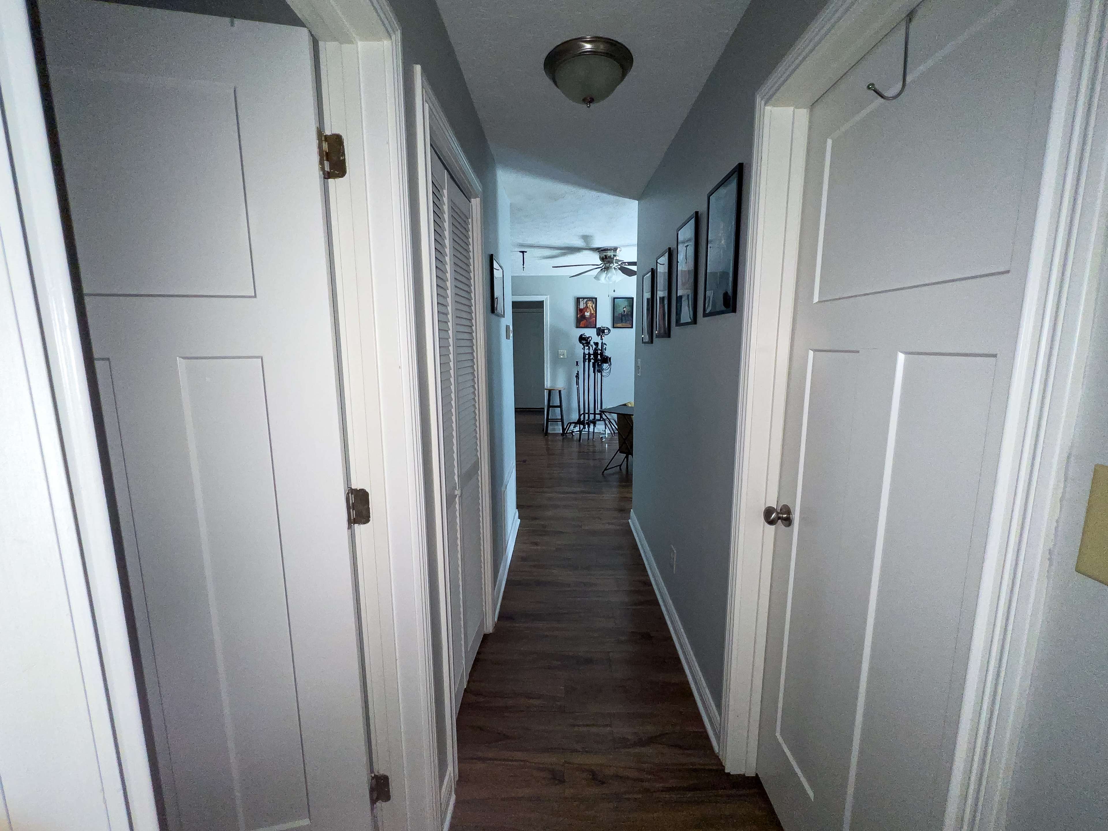 A narrow hallway with white doors on either side leads to a room at the far end, illuminated by overhead lighting.