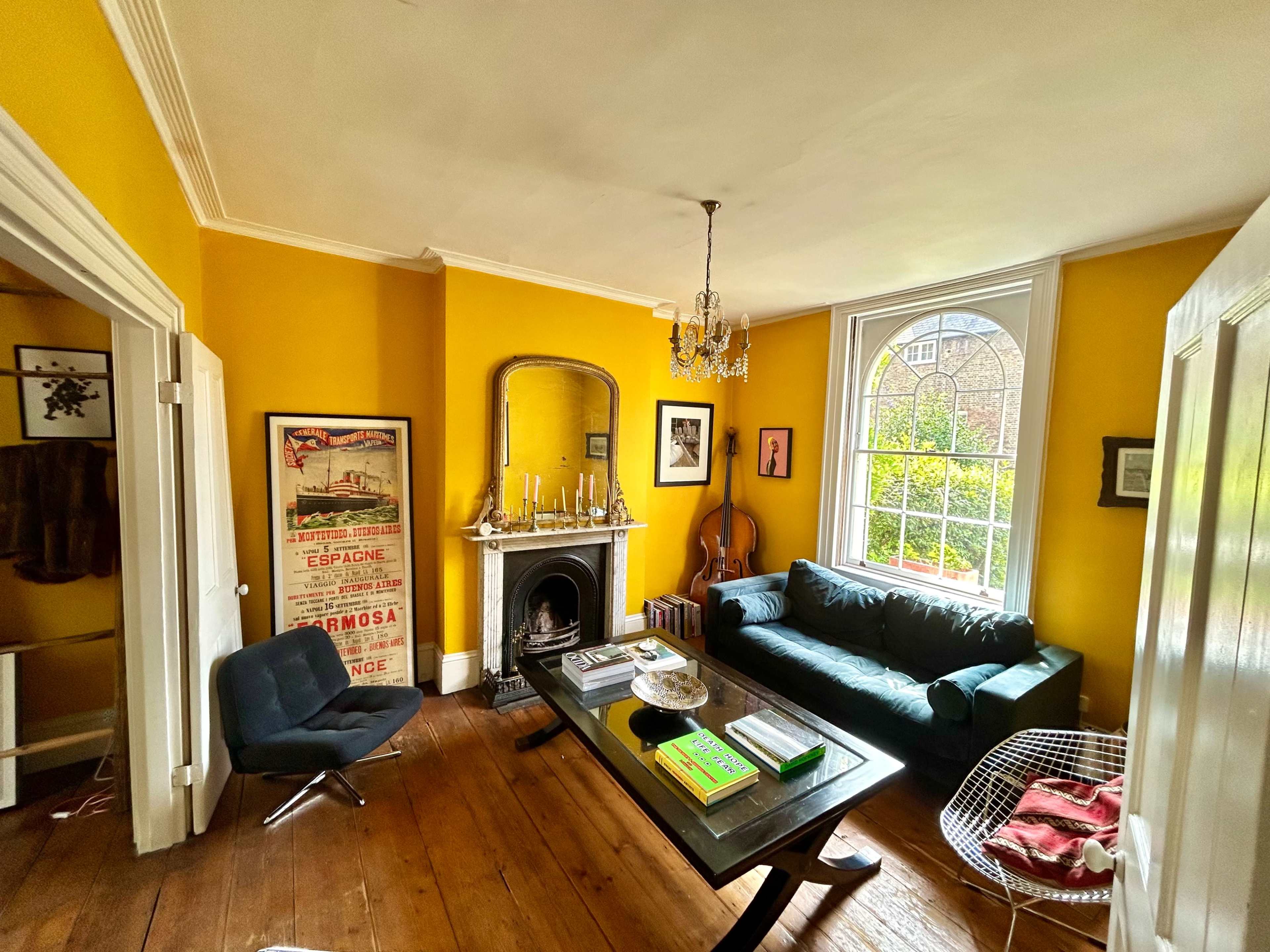 The image shows a cozy living room with yellow walls, a dark green couch, a coffee table, and a large window letting in natural light.