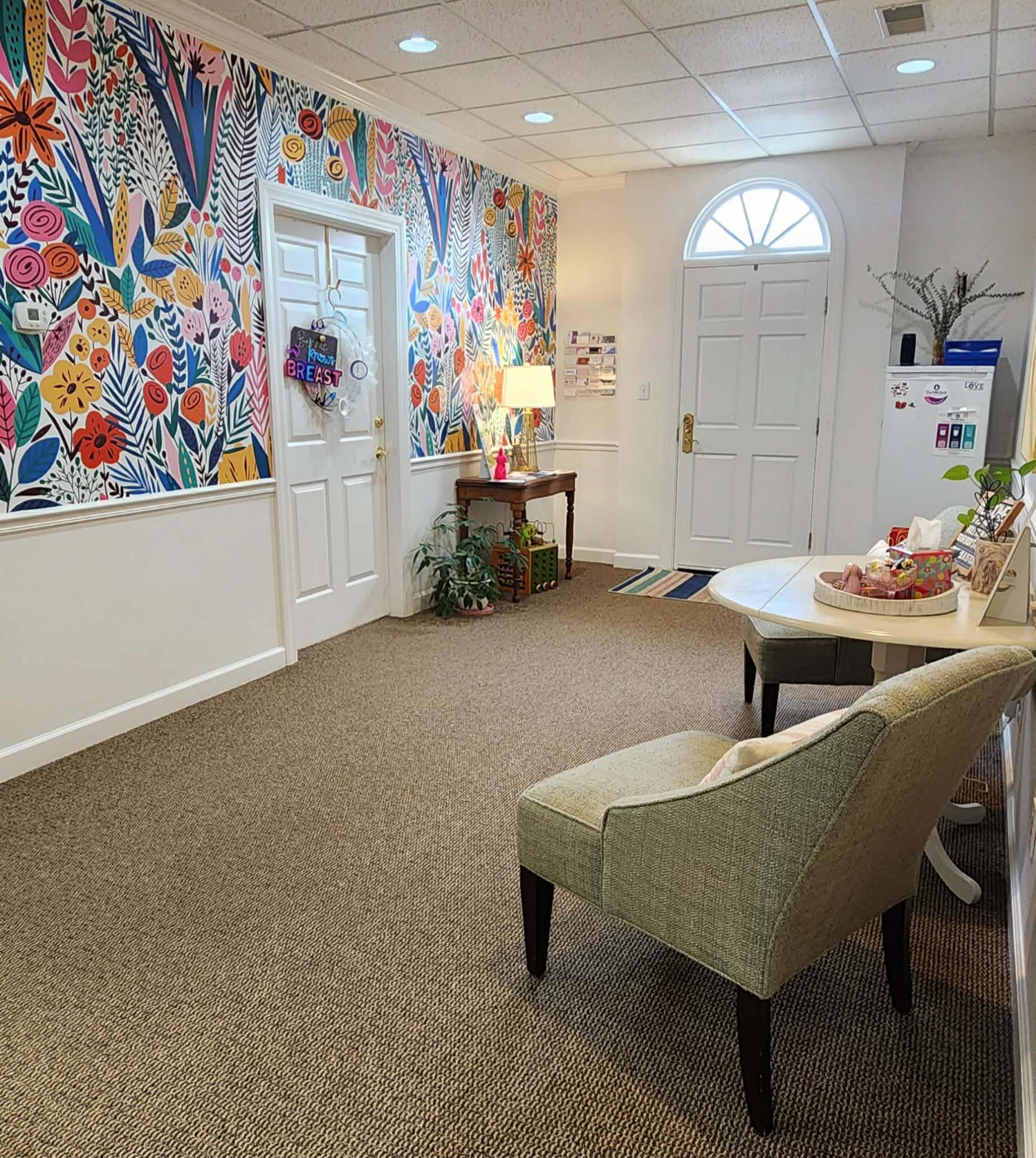 The image shows a brightly decorated lobby with floral wallpaper, a small table with a lamp, a chair, and a refrigerator against one wall.