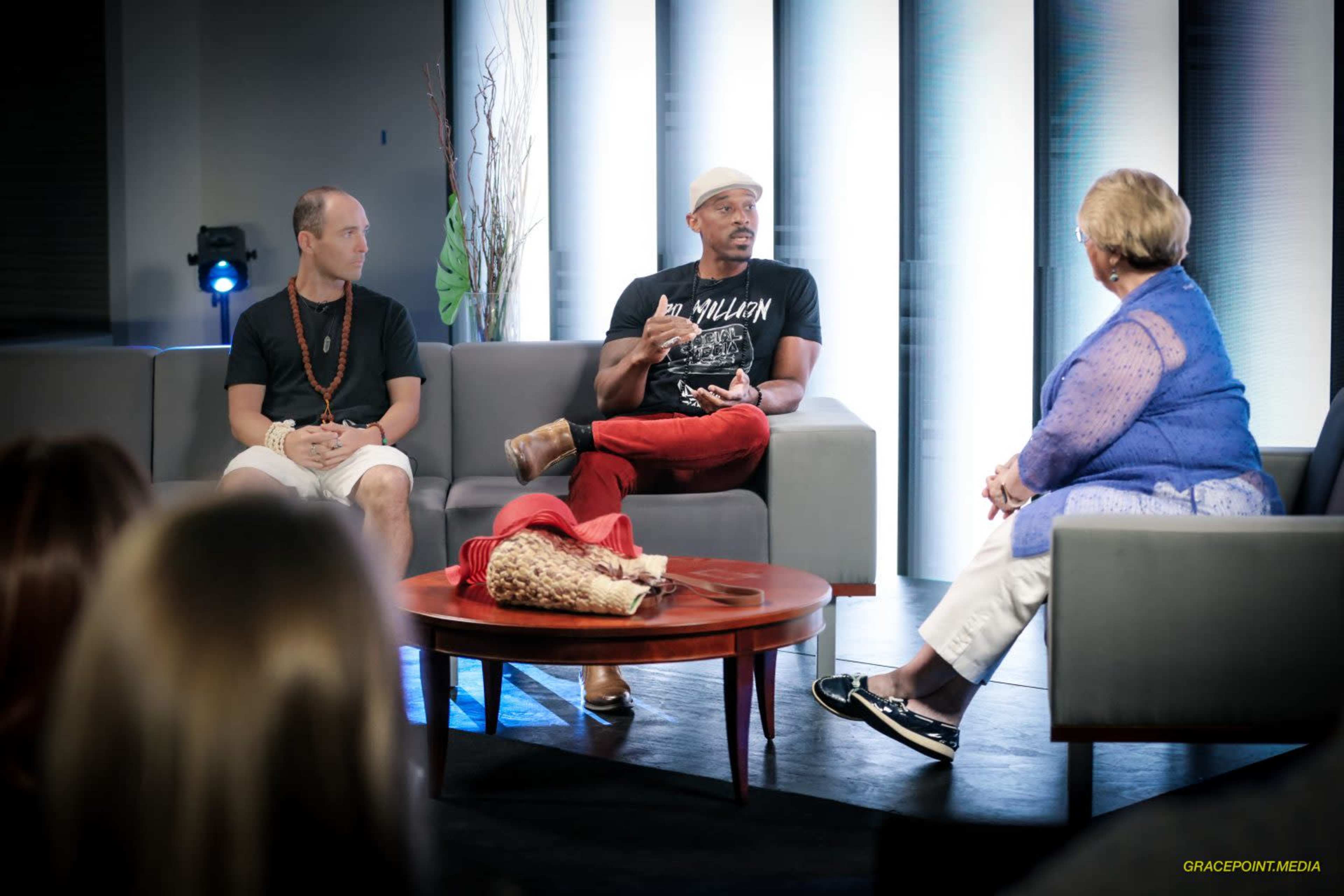 Three individuals are discussing on a stage with a circular table, surrounded by modern seating and a backdrop of lit panels.
