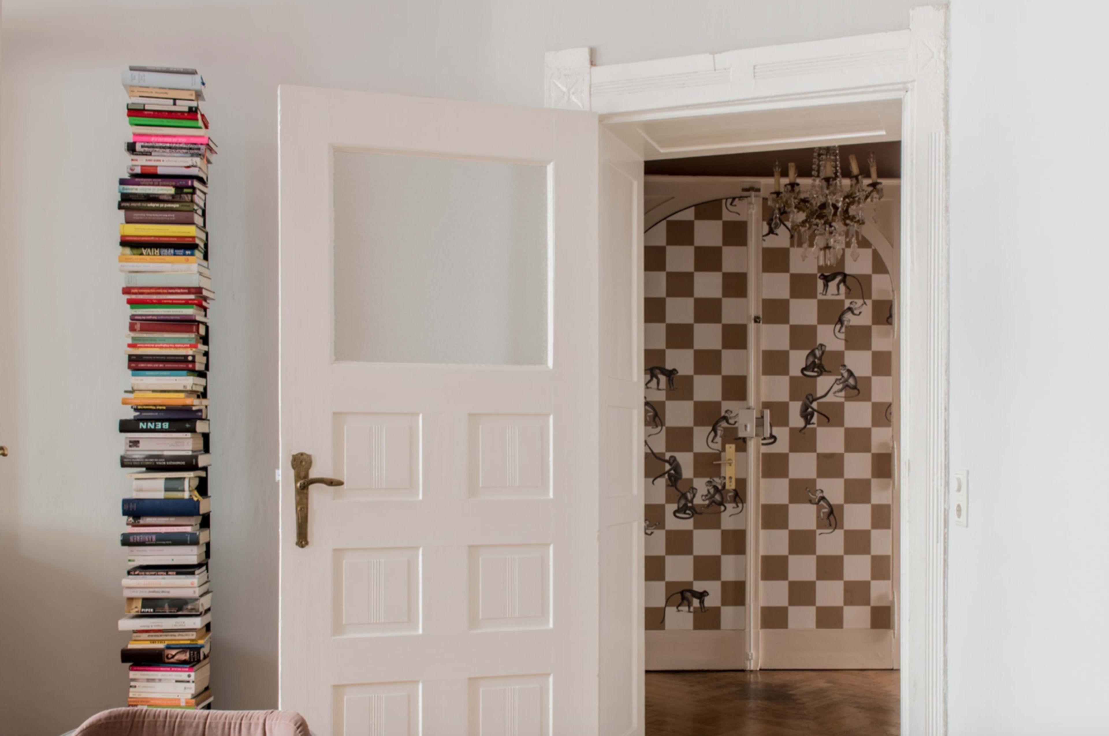 A white door stands open to reveal a patterned wall and a chandelier, with a tall stack of books beside the entrance.