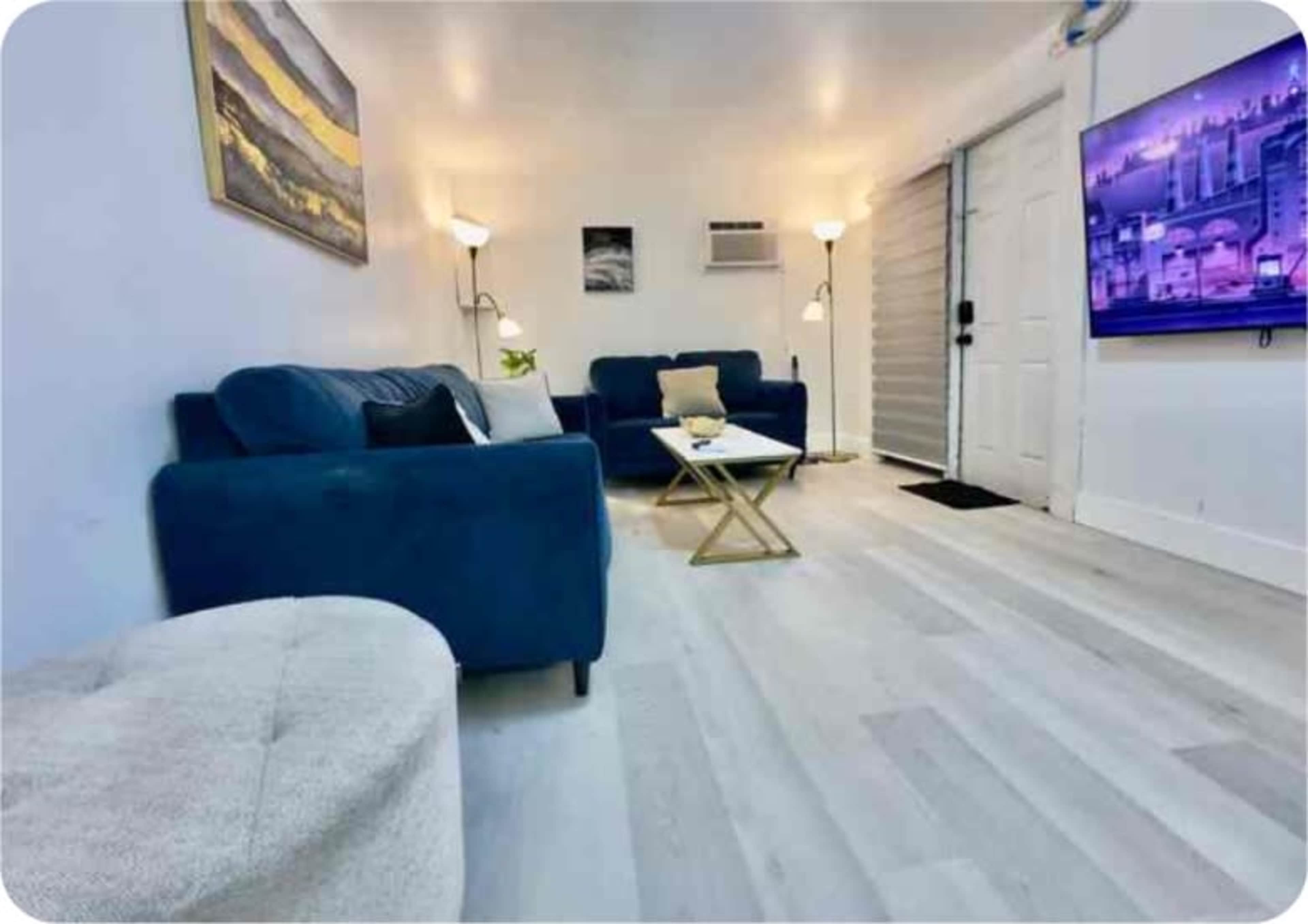 The image shows a modern living room featuring a blue couch, a light-colored ottoman, a small coffee table, and a television mounted on the wall.