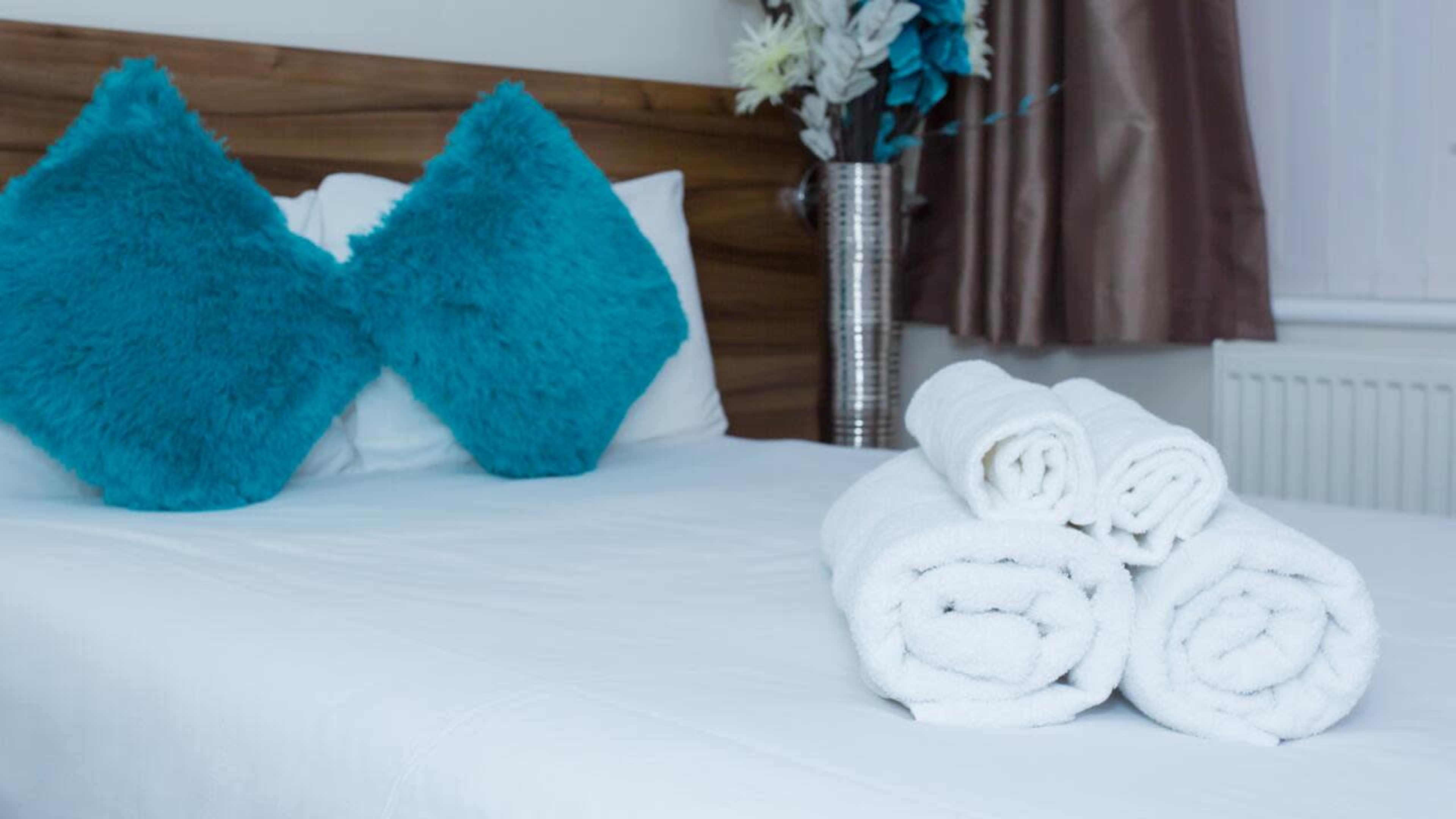A neatly made bed with white linens features rolled white towels and turquoise decorative pillows.