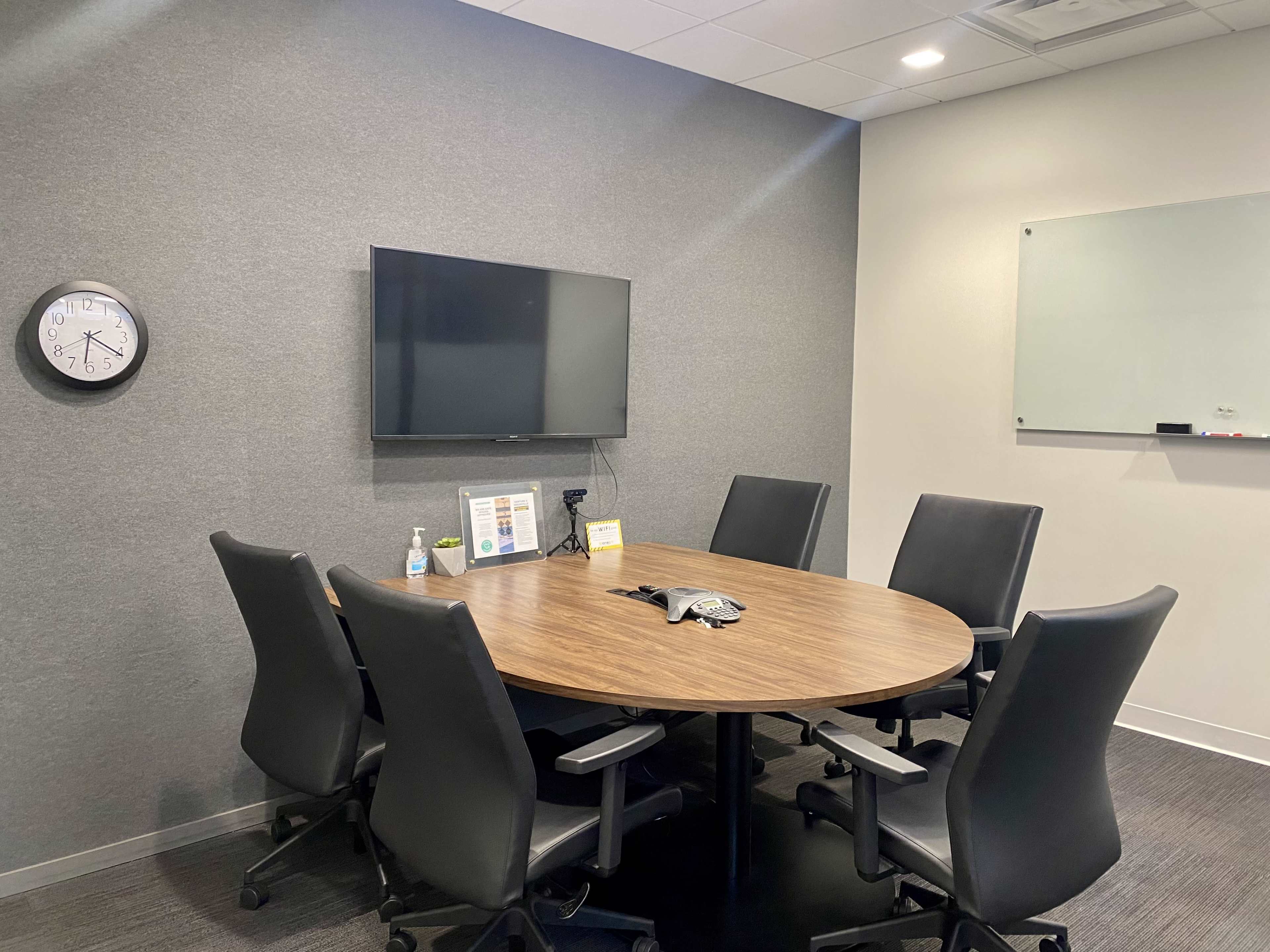 A modern conference room with a round wooden table surrounded by six black ergonomic chairs, a wall-mounted TV, a clock, and a whiteboard.