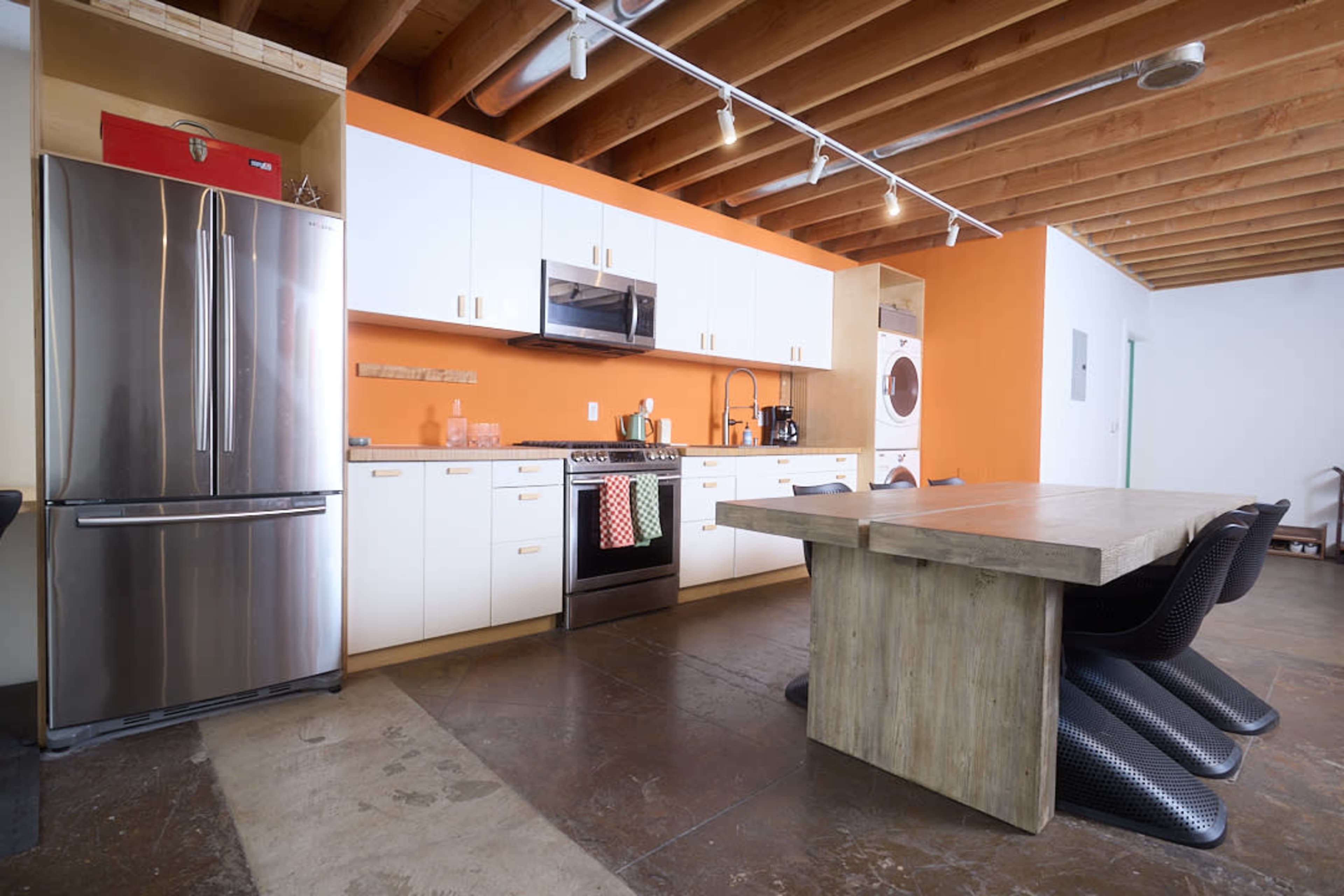 The image shows a modern kitchen featuring stainless steel appliances, white cabinetry, an orange accent wall, and a wooden dining table with black chairs.
