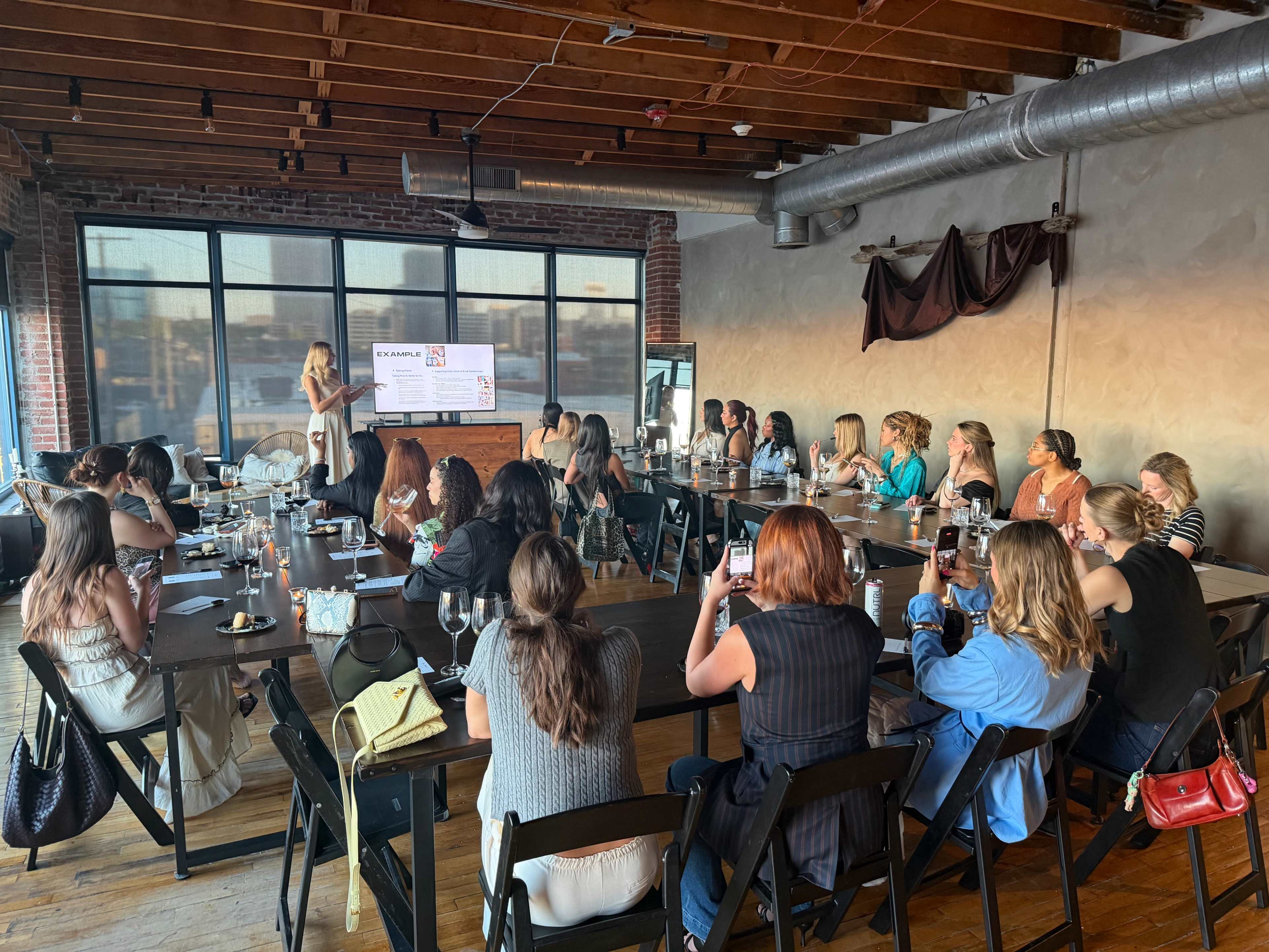 A group of people is seated at a long table in a well-lit room with large windows, attentively watching a presentation being given at the front.