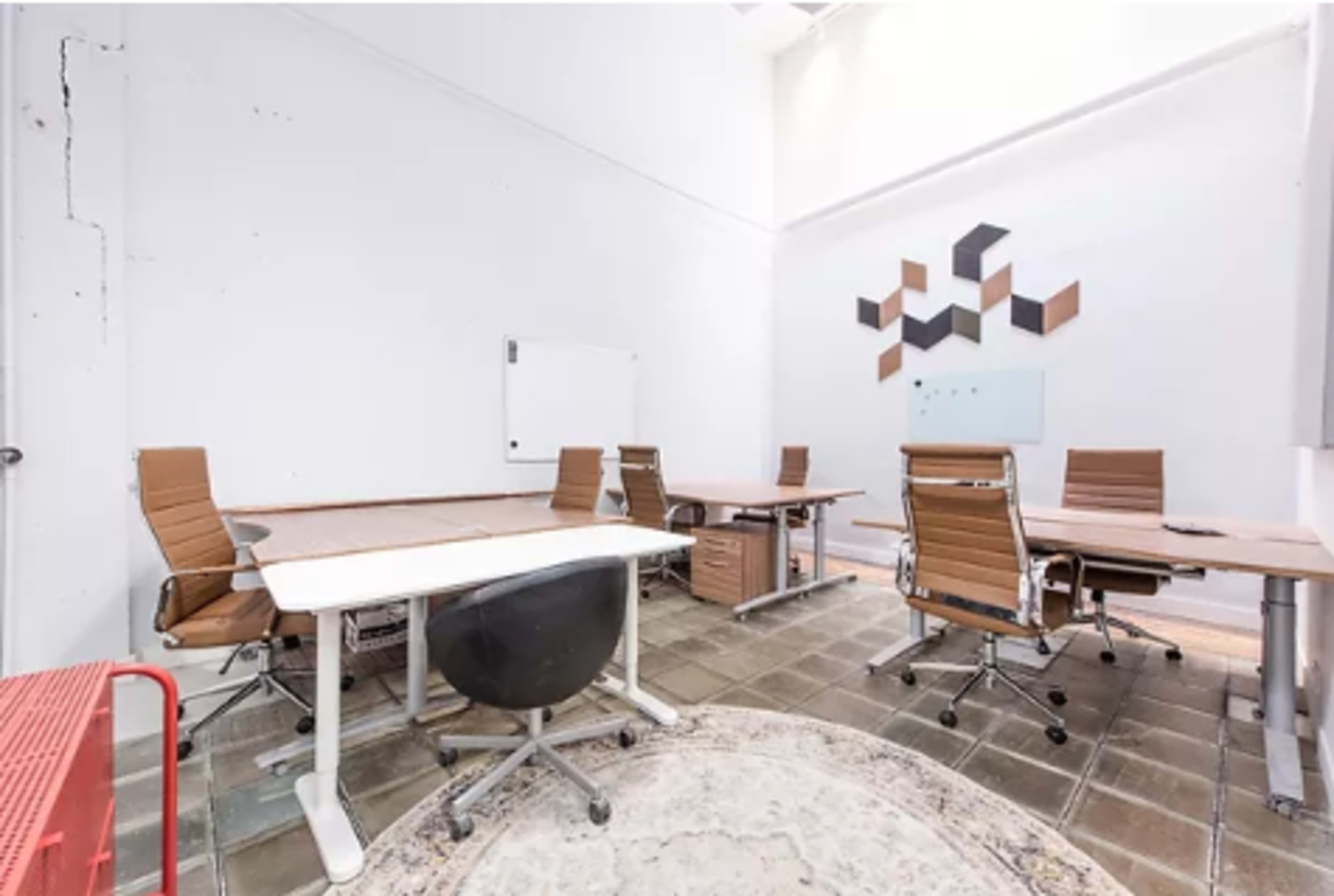 A bright, modern office space features multiple desks with ergonomic chairs, a whiteboard, and geometric wall decor.