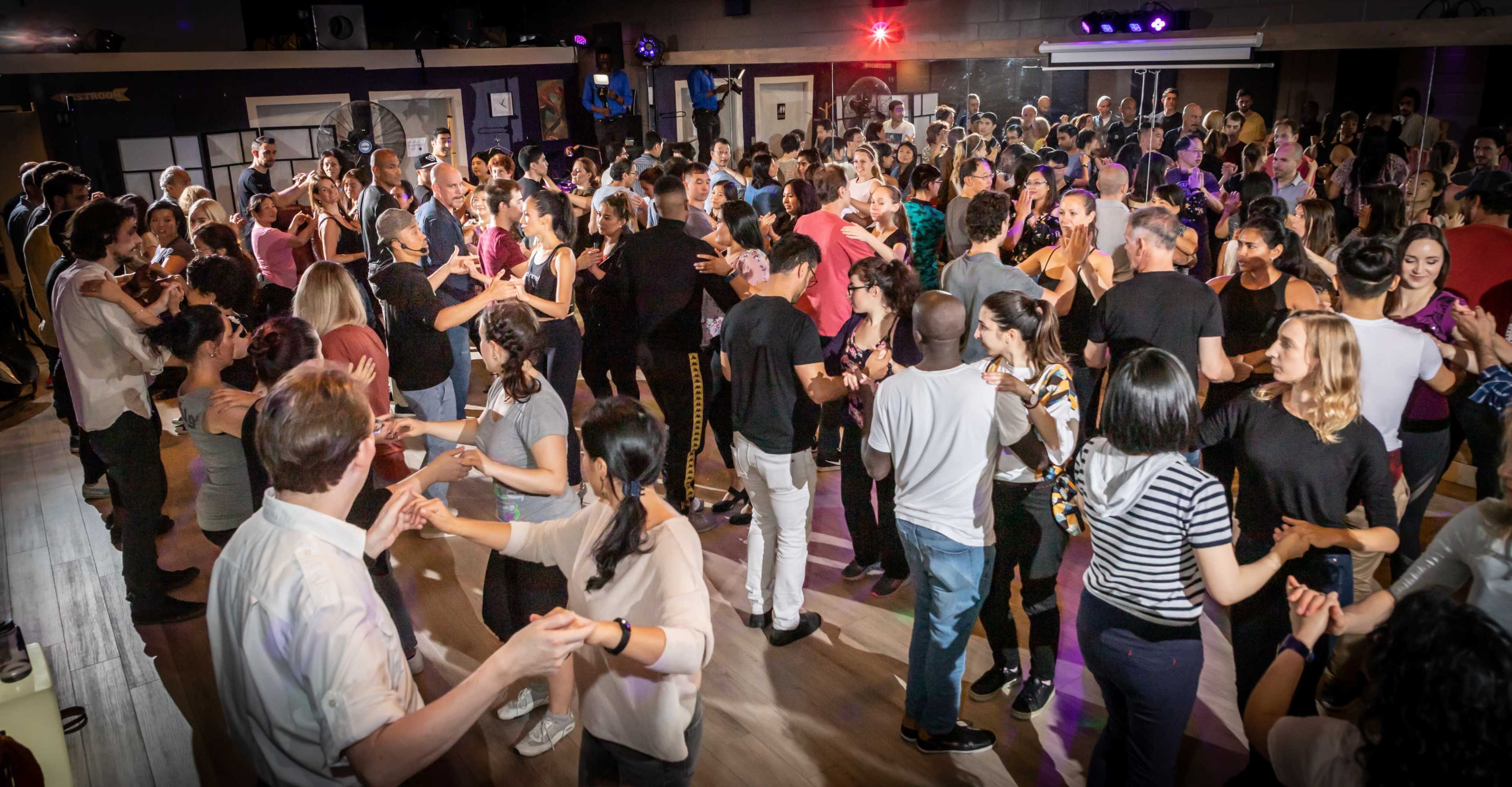A large group of people dance together in a bustling dance studio with colorful lights and a lively atmosphere.