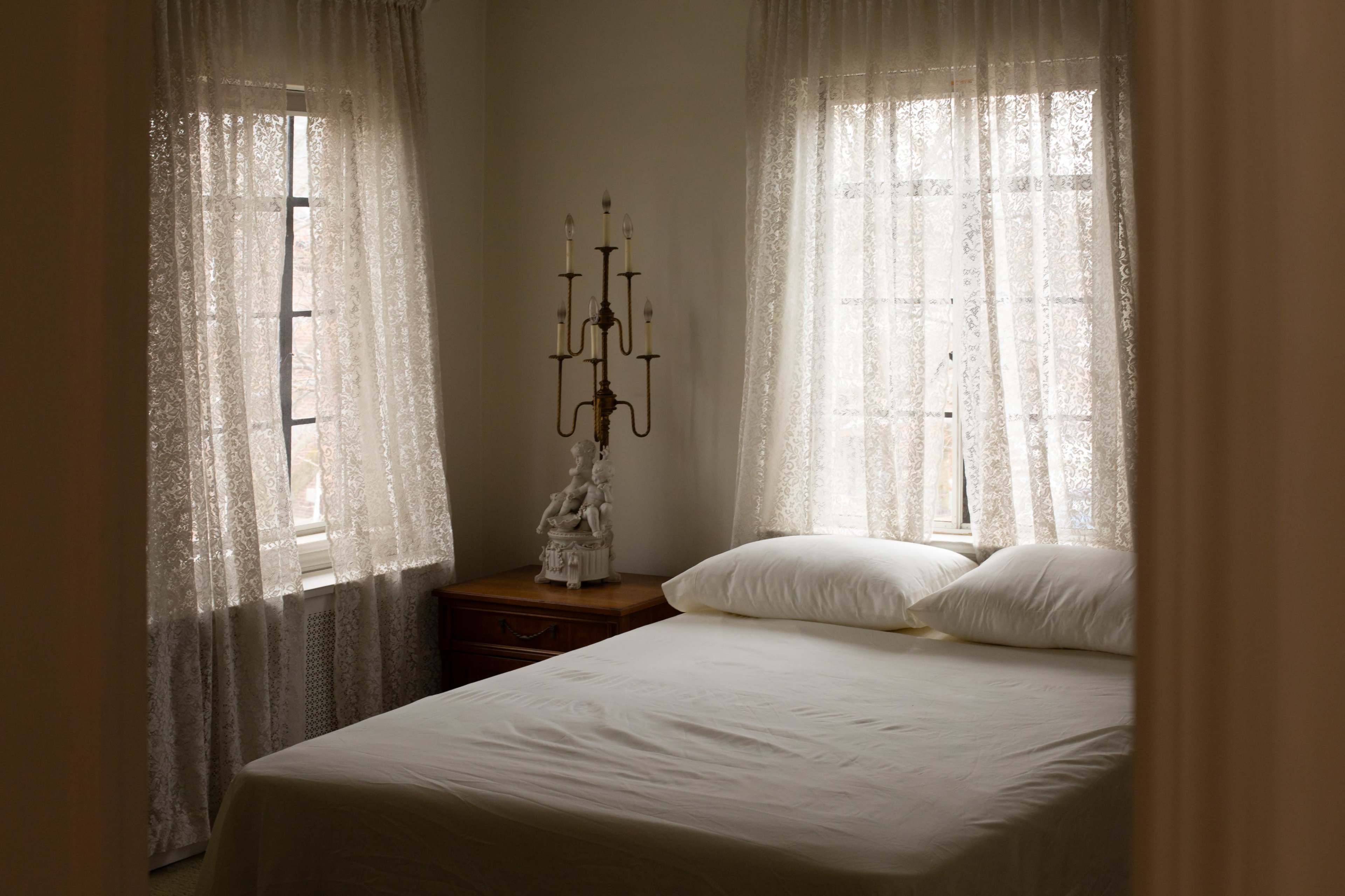 The image shows a softly lit bedroom featuring a neatly made bed with white linens, a wooden nightstand, and sheer, lace curtains framing two windows.