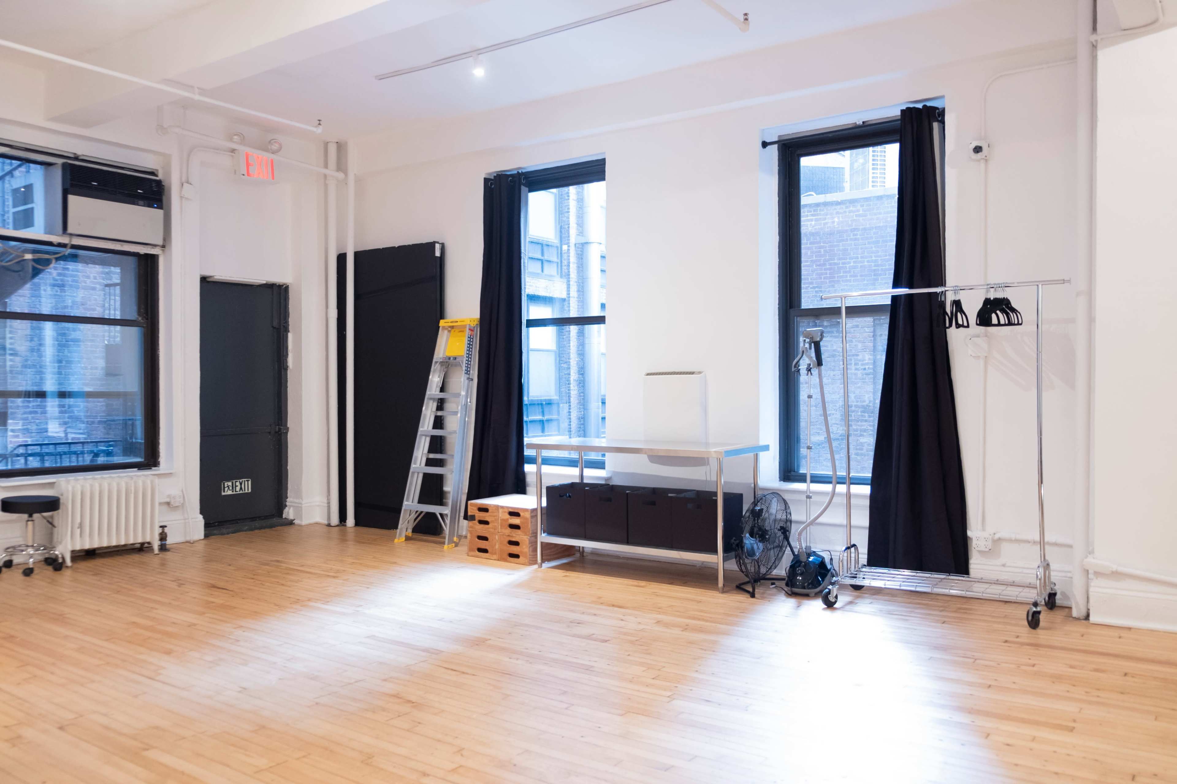 The image shows a spacious, well-lit room with two large windows, a ladder, a table, a clothing rack, and wooden flooring.