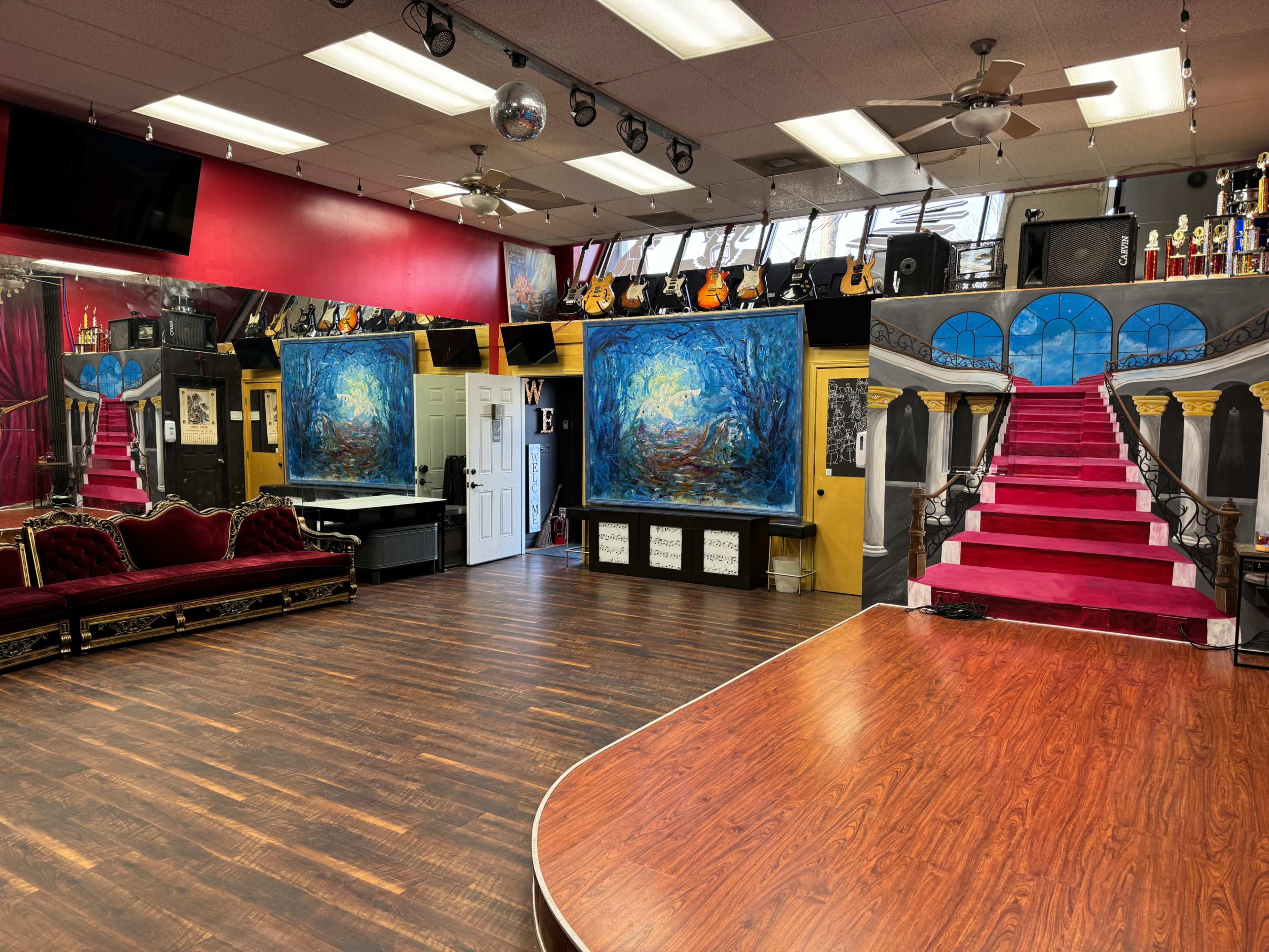 A dance studio with hardwood flooring, featuring a stage with red stairs, large wall paintings, and decorated with musical instruments and trophies.