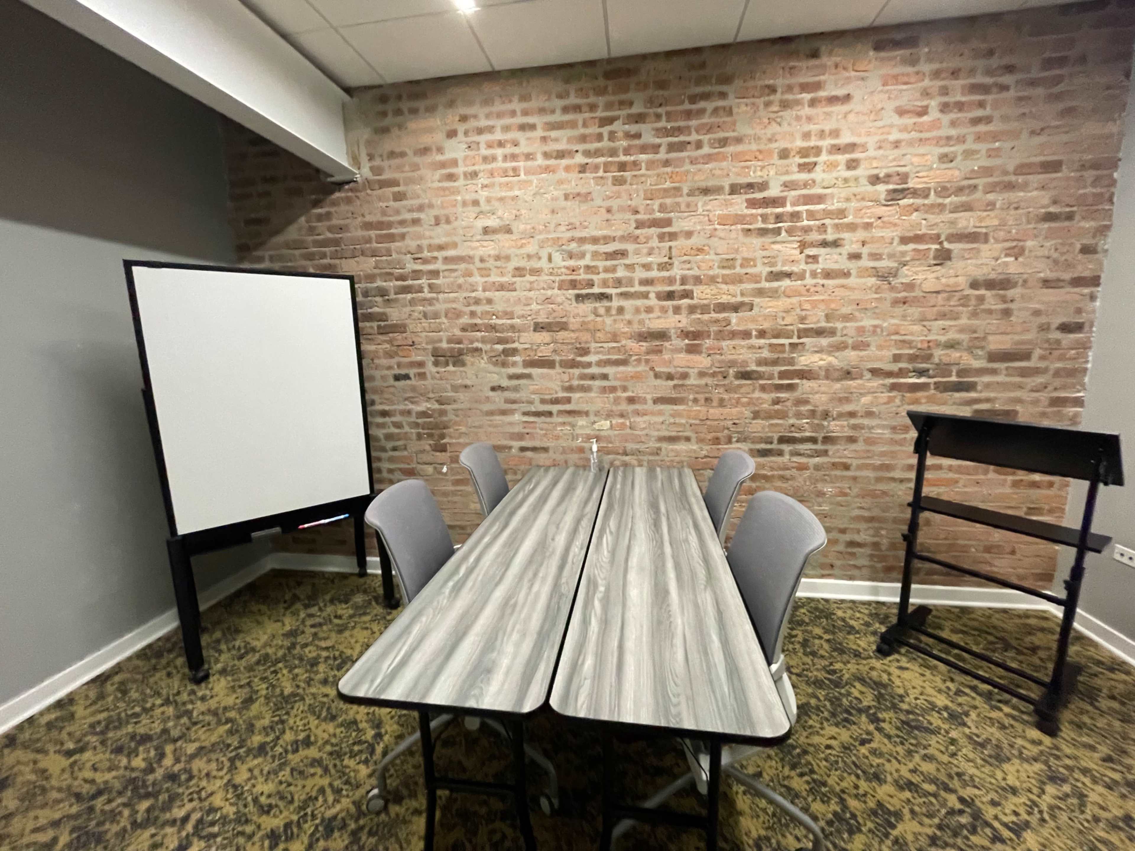 A small meeting room features a rectangular table with gray chairs, a whiteboard on a stand, and a wooden podium against a brick wall.