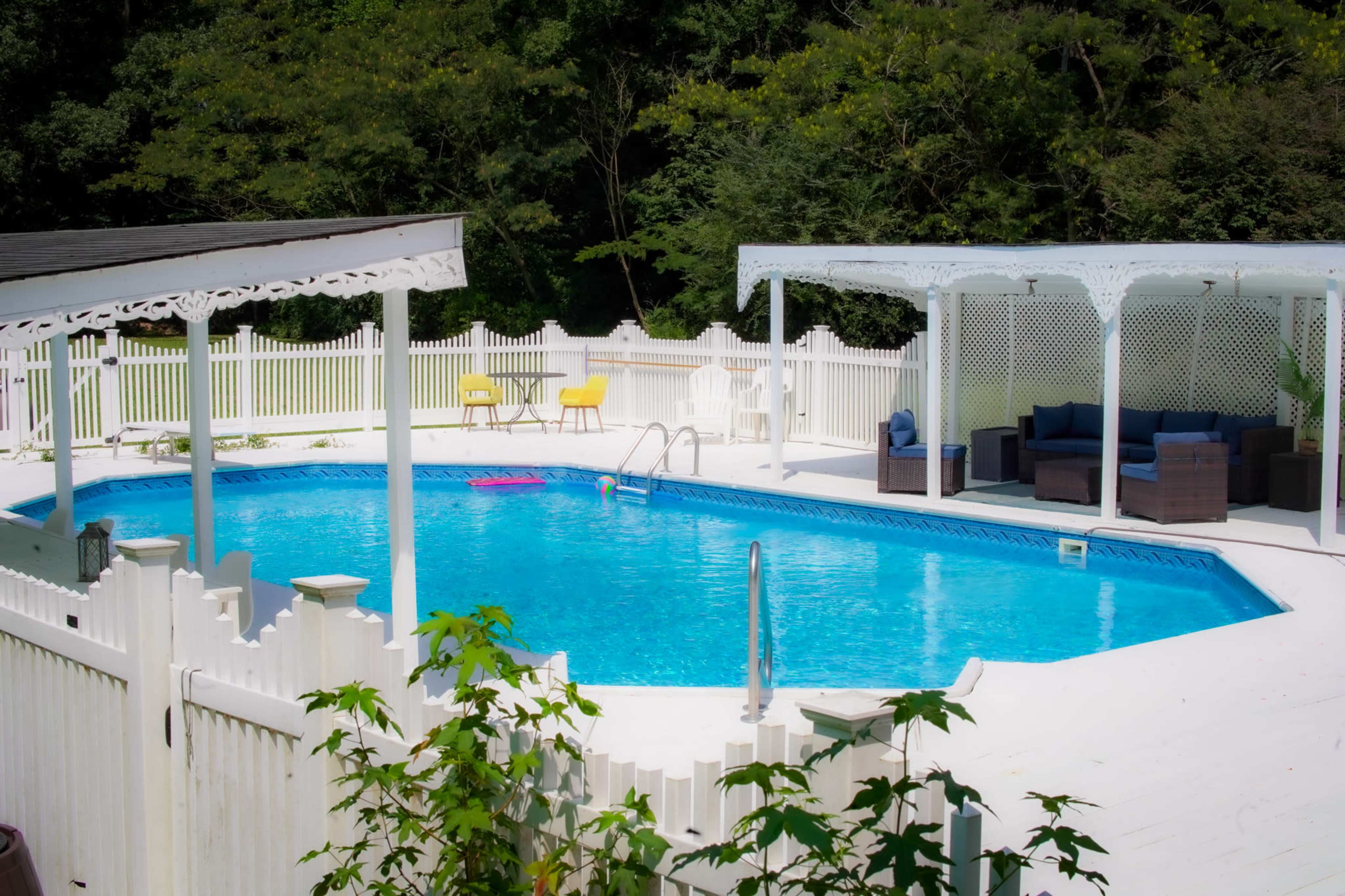 A fenced-in backyard with a rectangular swimming pool surrounded by lounge chairs and a covered seating area.