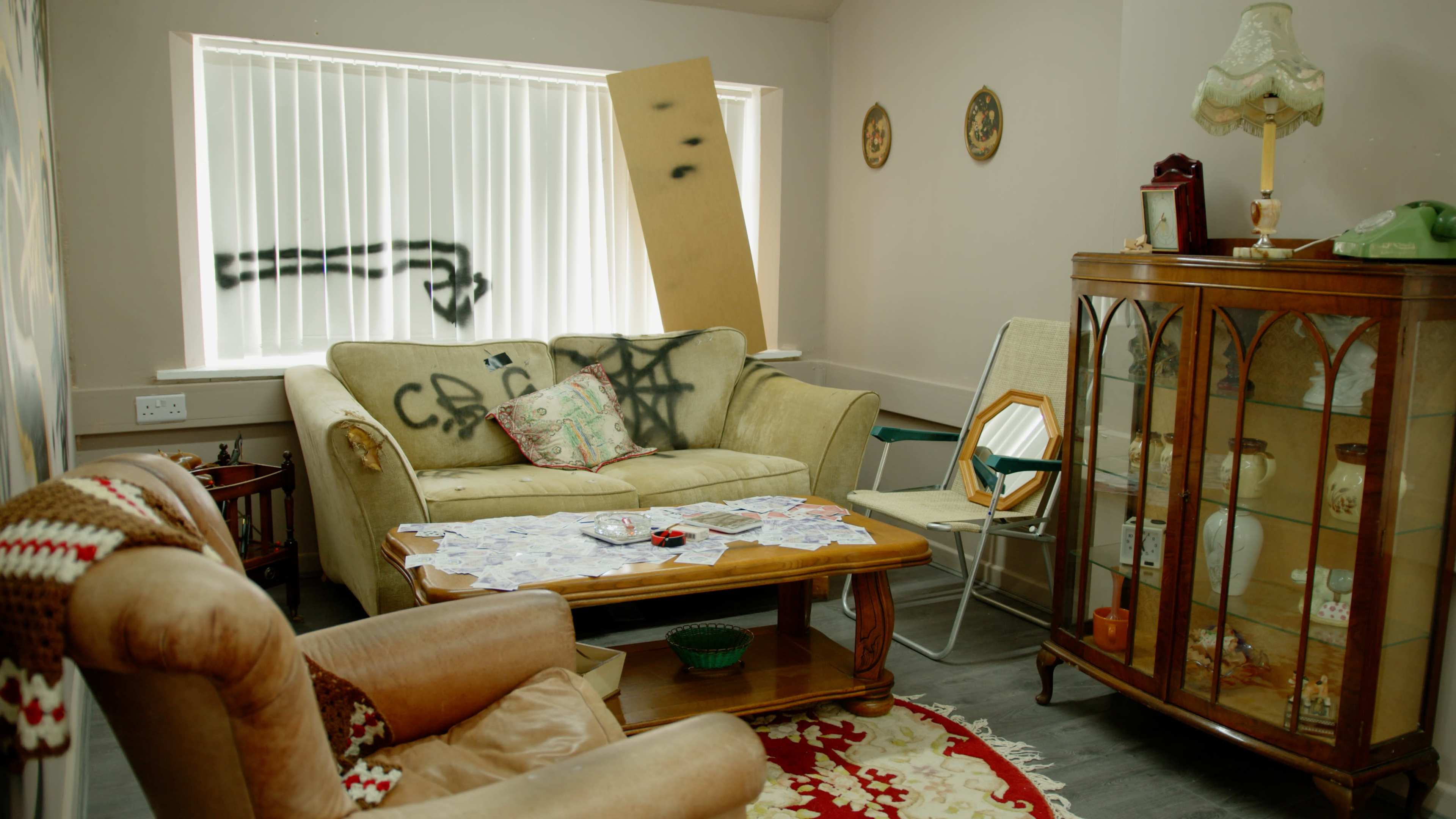 The scene shows a cluttered living room with a sofa, a coffee table covered in papers, a wooden cabinet, and graffiti on the walls.