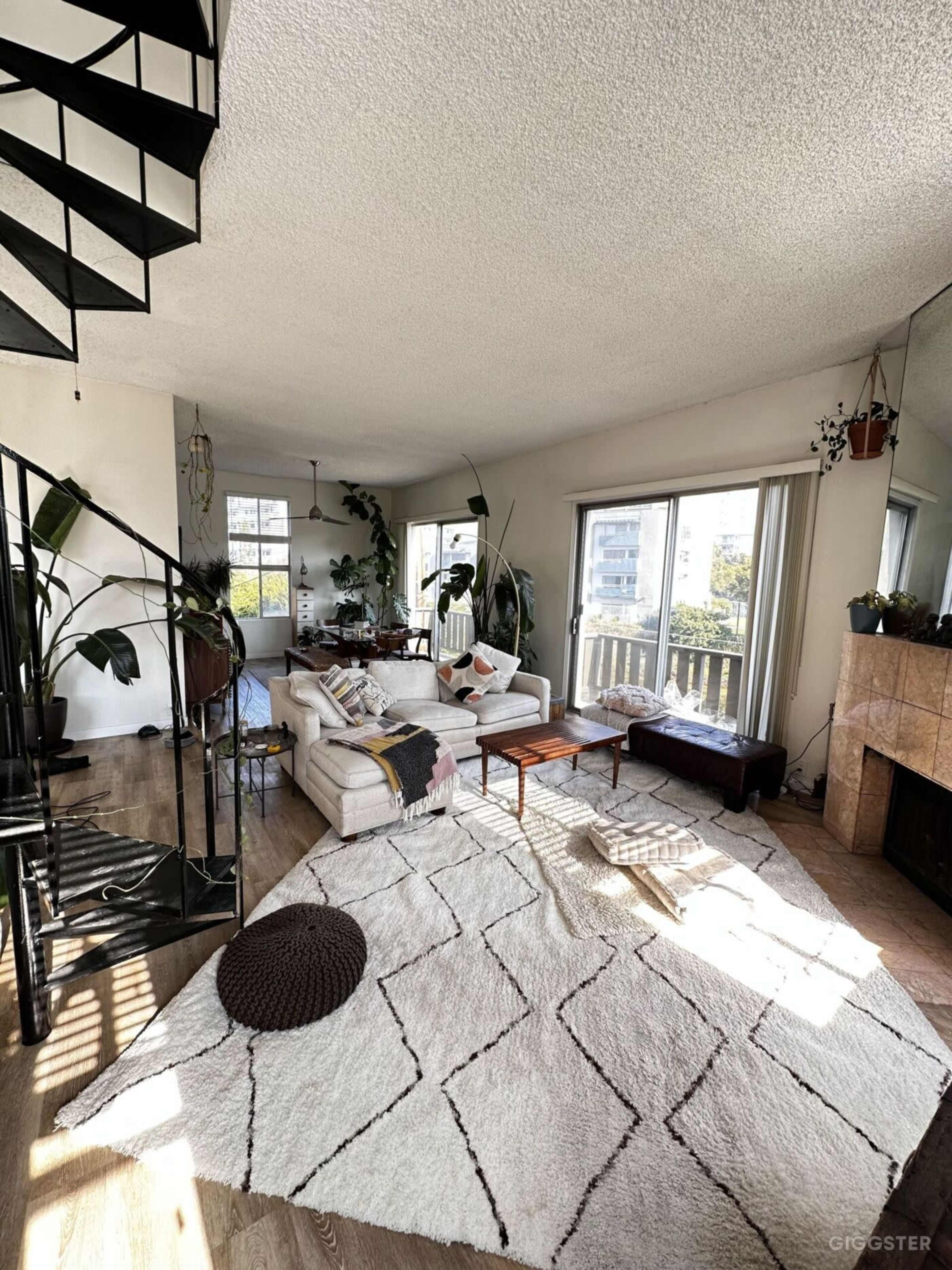 A spacious living room features a large sectional couch, a wooden coffee table, and a spiral staircase, surrounded by numerous plants and bright natural light.