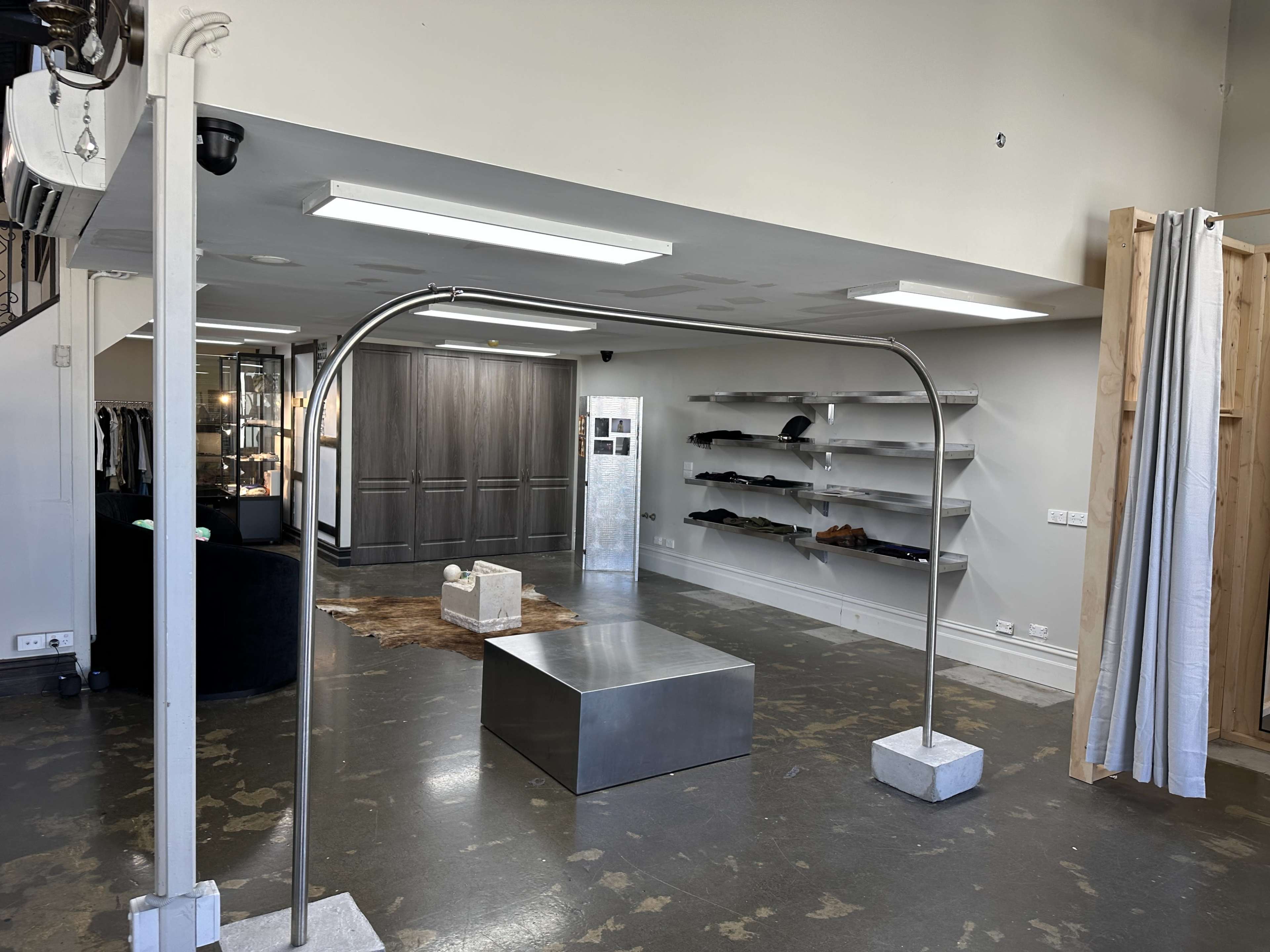 The image shows a modern retail space featuring a central metallic display table, clothing racks on the walls, and a minimalist aesthetic with concrete flooring.