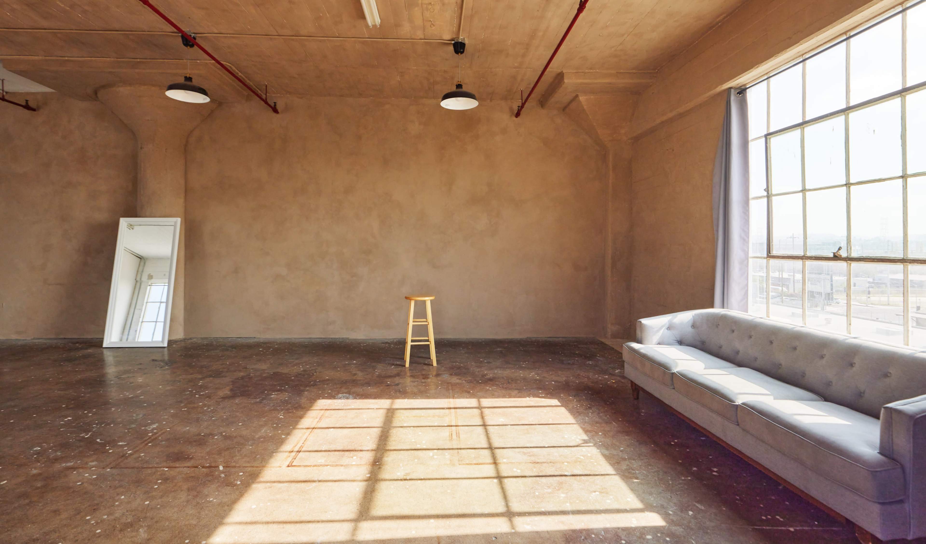 A spacious, empty room features a wooden stool and a gray couch, illuminated by sunlight streaming through a large window.