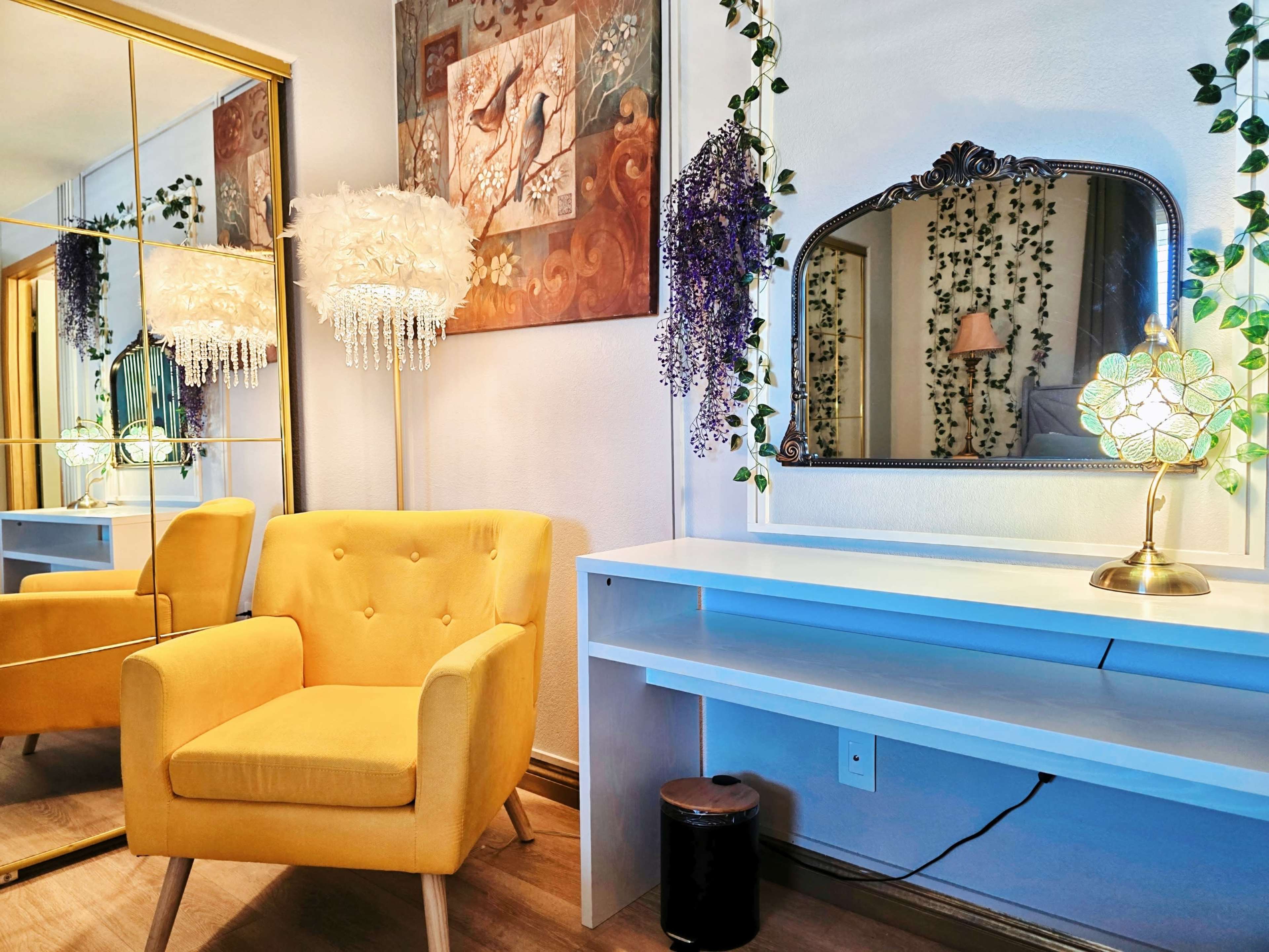 The image shows a bright yellow armchair next to a mirrored wall and a dressing table with a decorative lamp, surrounded by indoor plants and artwork.