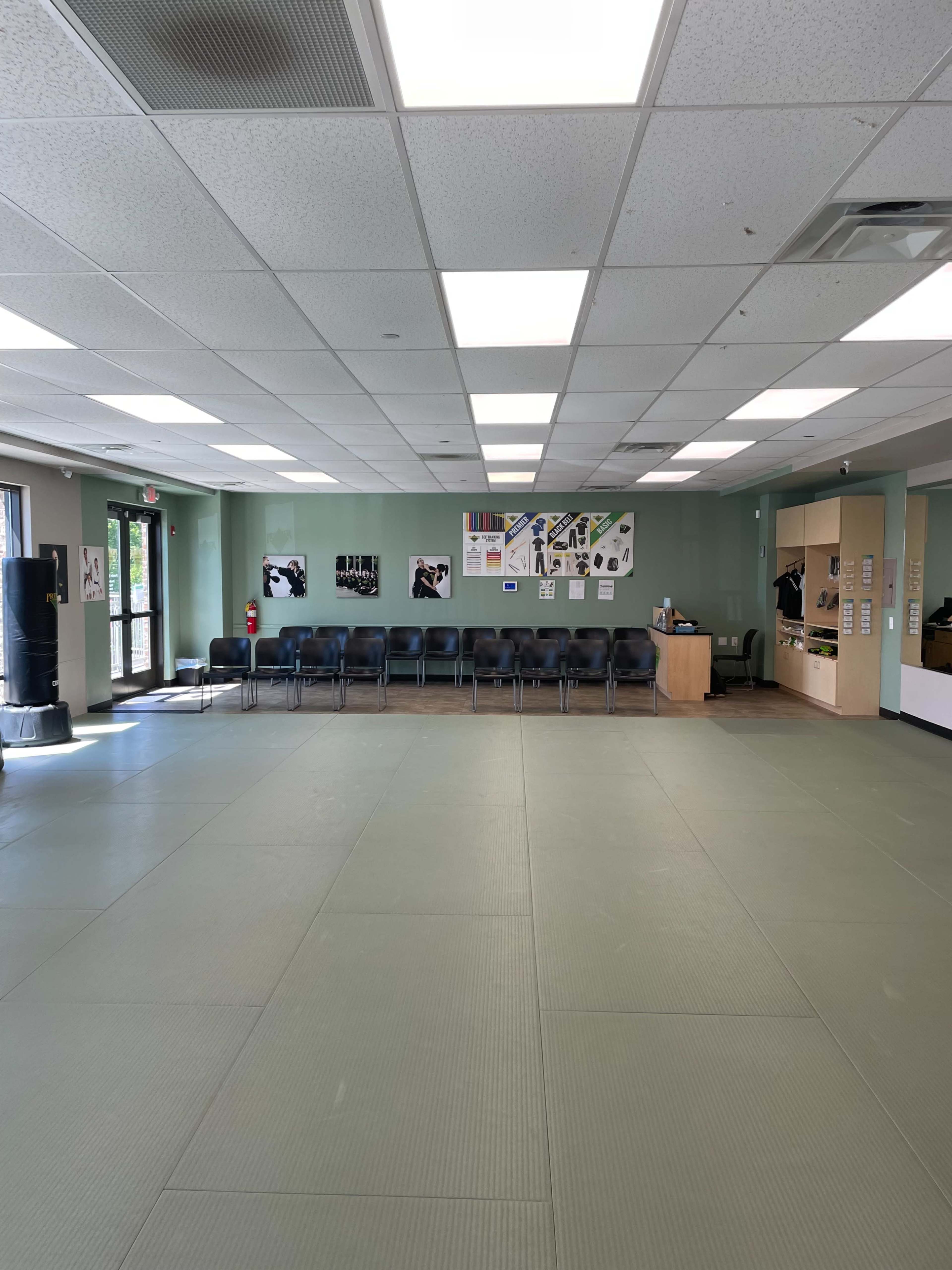 The image shows a spacious martial arts studio with a green mat floor, a row of black chairs along one wall, and a reception area with posters and informational materials.