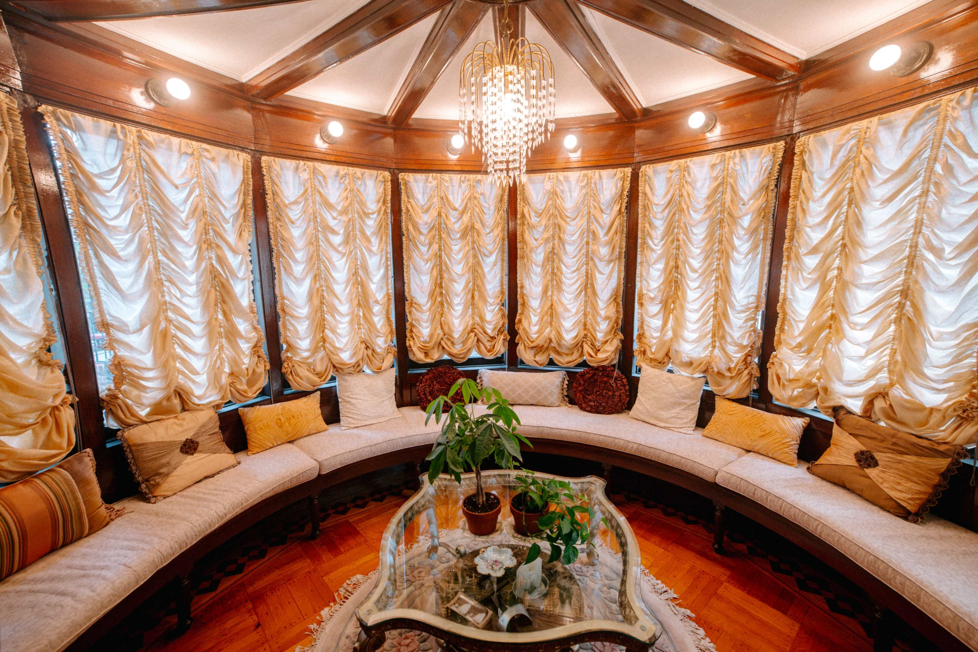 The image shows a circular room with a curved seating area, large decorative curtains, and a glass coffee table featuring potted plants.